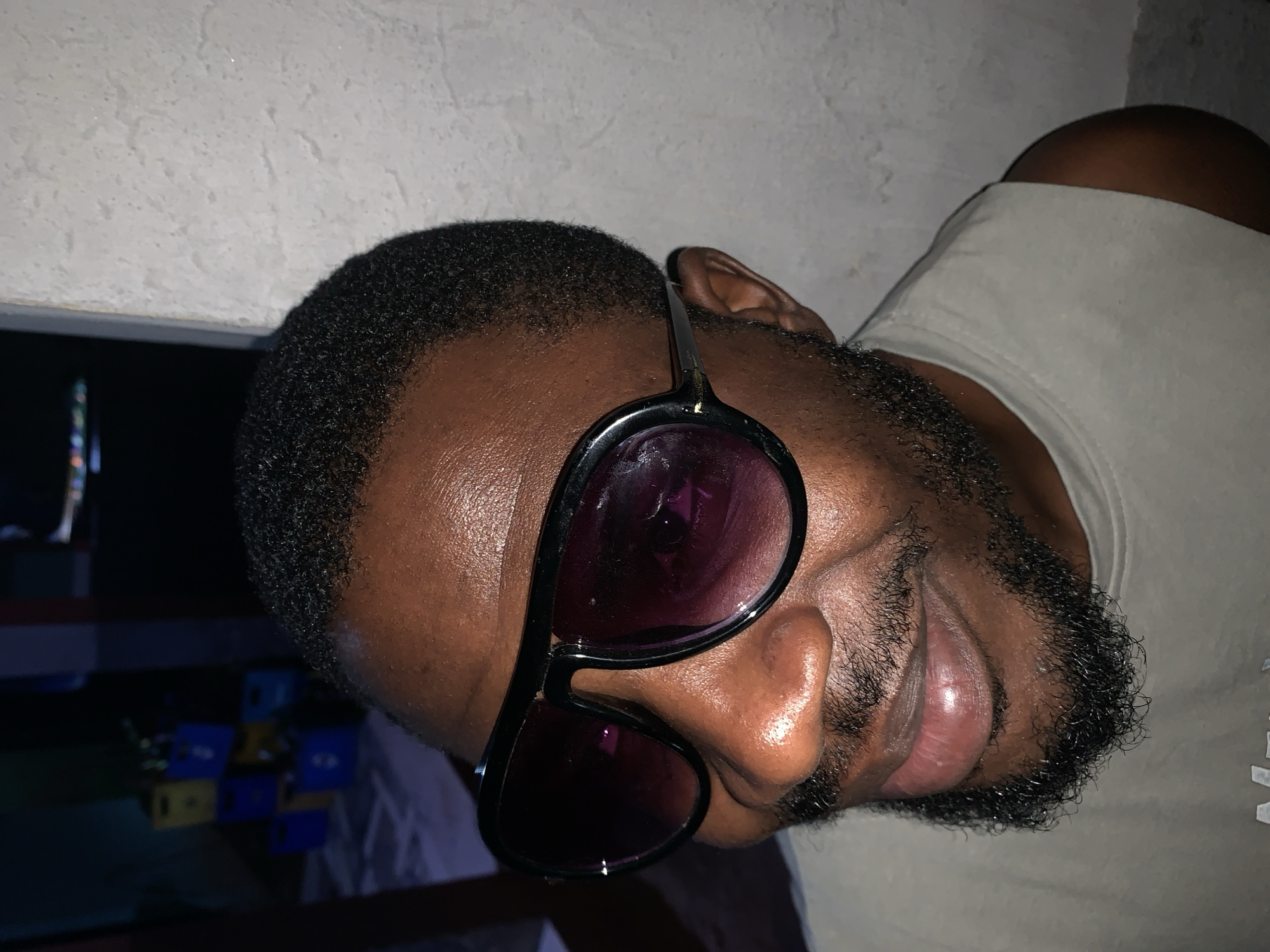 A close-up, slightly rotated photograph shows a man with dark skin, short black hair, and a full beard. He is wearing sunglasses with dark lenses and black frames. He is wearing a light-colored t-shirt. The background is blurry, but there appears to be a light-colored textured wall behind him, and other objects in the distance. The photograph appears to be taken indoors. The image contains no information about the city of New Stadium, Nigeria.