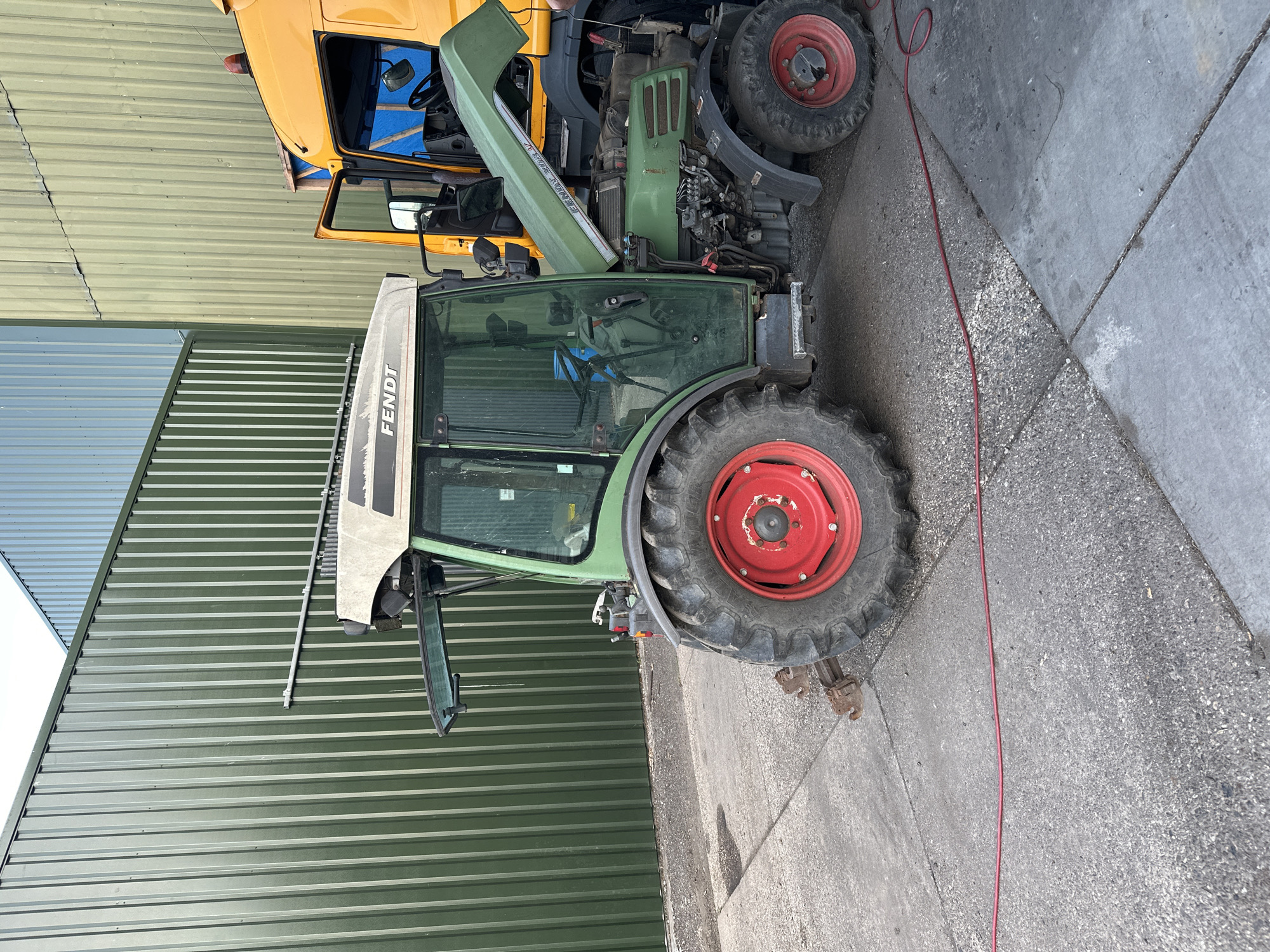 An agricultural Fendt 208 V tractor with its hood open is parked on a concrete surface next to a large yellow truck. The tractor is green with red rims on its wheels. The truck is positioned to the right of the tractor, and its driver's side door is open. The scene appears to be an outdoor industrial or agricultural setting, likely a farm or a service yard. The background consists of a tall, ribbed green metal building and a section of a light blue metal building. A red cable is visible running across the concrete in the foreground. The location is the city of Dronten, Netherlands.