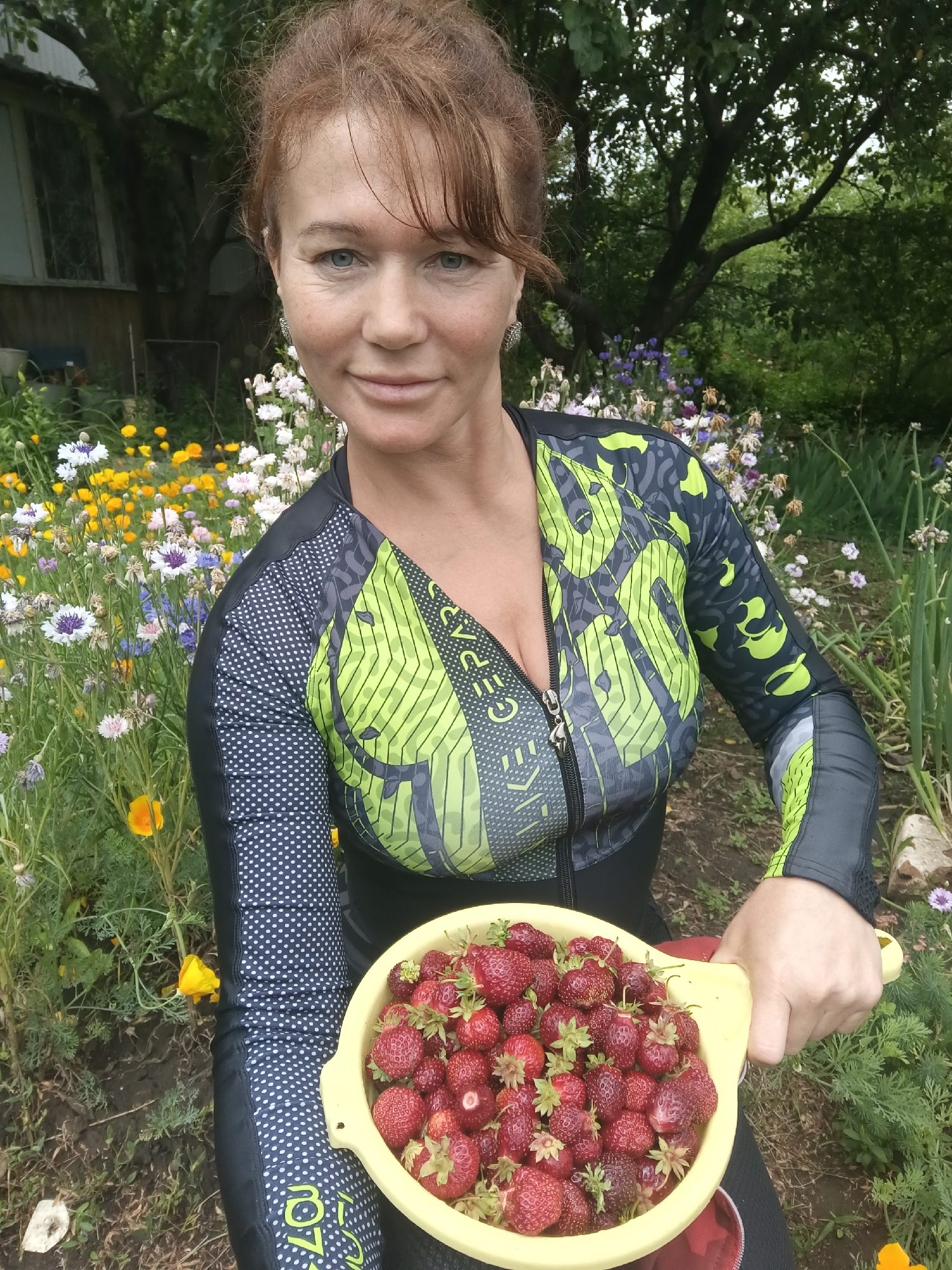 In the village of Vyazovka, Russia, a woman with auburn hair and light blue eyes is pictured outdoors. She is wearing a long-sleeved, black and neon green cycling jersey. In her hands, she holds a yellow bowl filled with freshly picked strawberries. The background consists of a lush garden with a variety of flowers, including yellow poppies and purple and white cornflowers, as well as a glimpse of a wooden house. The scene appears to be set in a rural environment during daylight hours.