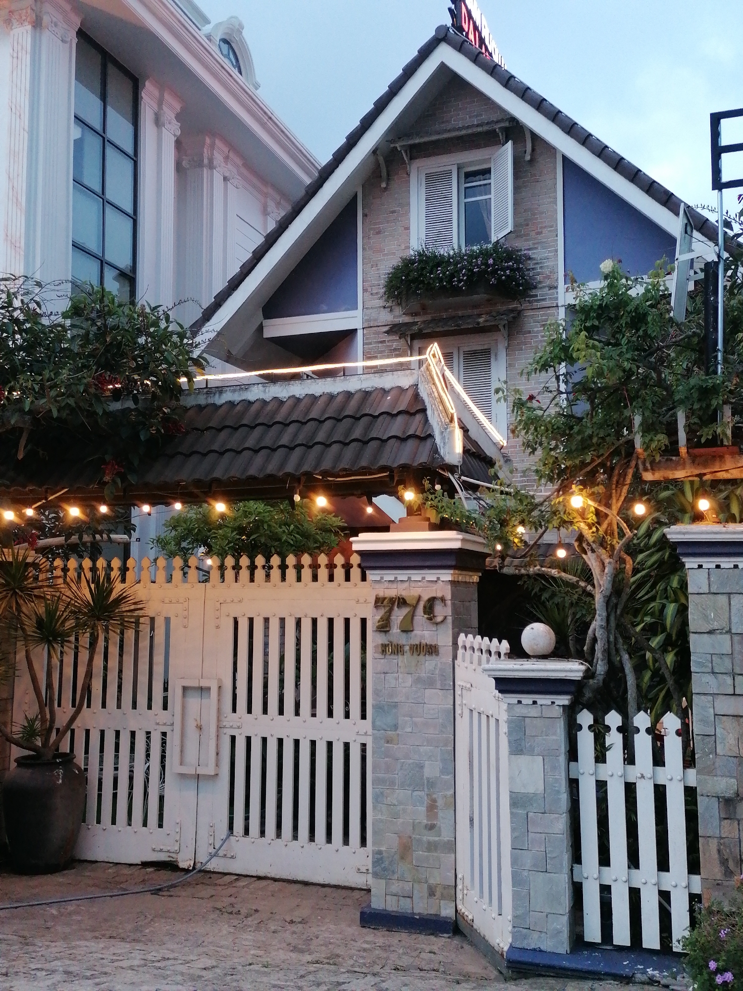 The image shows a residential property entrance with a white metal gate and a stone pillar in the city of Xuân Trường - Đà Lạt, Vietnam. The gate is ornate with vertical slats and decorative bolts. A stone pillar to the right of the gate has the number "77c" and text that appears to be "HUNG VUONG" written on it. The property is surrounded by lush greenery and trees, some of which are adorned with string lights. A large dark-colored ceramic pot containing a palm-like plant sits to the left of the gate. In the background, two different architectural styles of buildings are visible: a modern white building with classical columns and a brick house with a pitched roof. The sky is overcast, indicating either early morning or late evening. The scene appears to be well-maintained and evokes a sense of tranquility and elegance.