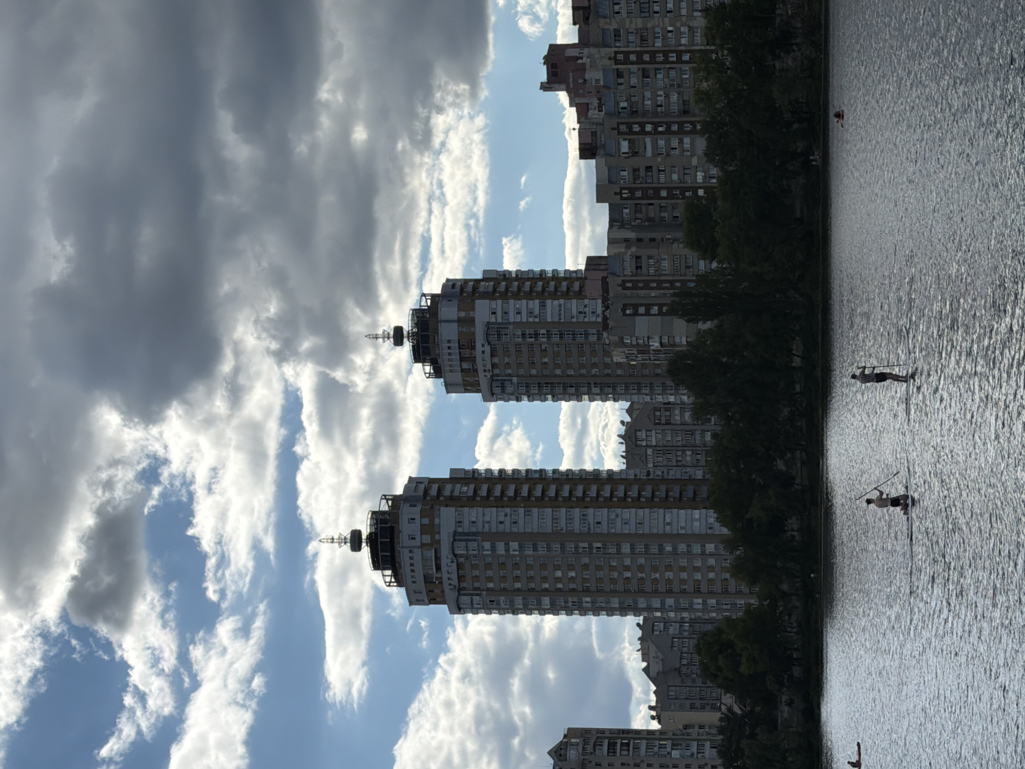 This is a wide shot of the Obolonska Naberezhna embankment in Kyiv, Ukraine, on a cloudy day. In the foreground, a wide body of water, likely the Dnipro River, reflects the sky and surrounding buildings. Three people are visible on the water. Two are on stand-up paddleboards, one of whom is kneeling and holding a paddle. Another person is on a small kayak or inflatable raft to the left.
In the background, a line of modern apartment buildings stretches across the horizon, with two distinctive tall, slender towers with antenna-like structures on top in the center of the frame. The buildings appear to be residential, with numerous windows and balconies visible. Dense green trees form a line between the water and the buildings. The sky is dramatic, with a mix of dark, heavy clouds and patches of bright blue sky. The overall scene depicts a recreational activity taking place in an urban environment with a prominent waterfront.