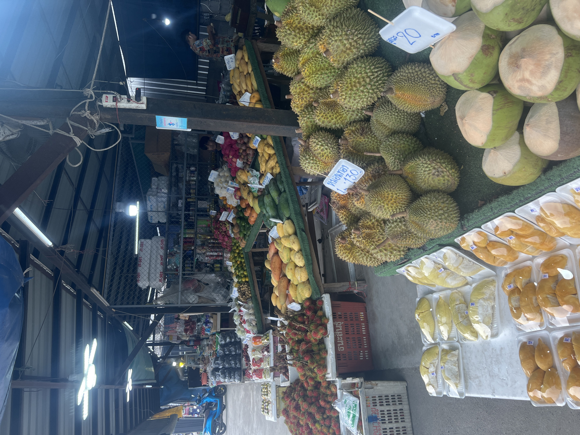 This is a photograph of an outdoor market stall in the city of Ko Samui District, Thailand, featuring a variety of fresh fruits. The stall is covered by a corrugated metal roof supported by wooden beams and poles, with blue tarps visible in the background. The ground is paved with concrete.
In the foreground, a display of whole durian fruits is arranged on the ground. To the left of the durians, there are several trays of pre-cut durian pieces, some wrapped in plastic. Further left, large piles of rambutans are displayed in red and white plastic crates. A blue scooter is parked to the far left, near a rack of plastic bags.
In the midground, various fruits are displayed on tiered tables. These include mangoes, papayas, and what appear to be avocados or similar green fruits. There are also piles of what seem to be lychees or similar small red fruits. Baskets and crates are used to hold and display the produce.
In the background, more produce is visible, along with shelves stocked with various items, including bottled drinks and packaged goods. Two individuals are present: a person in the midground to the left, partially obscured, appears to be attending to the fruit stall. Another person, wearing a patterned shirt, is visible in the background on the right, near the shelves. The overall impression is that of a busy, well-stocked local market.