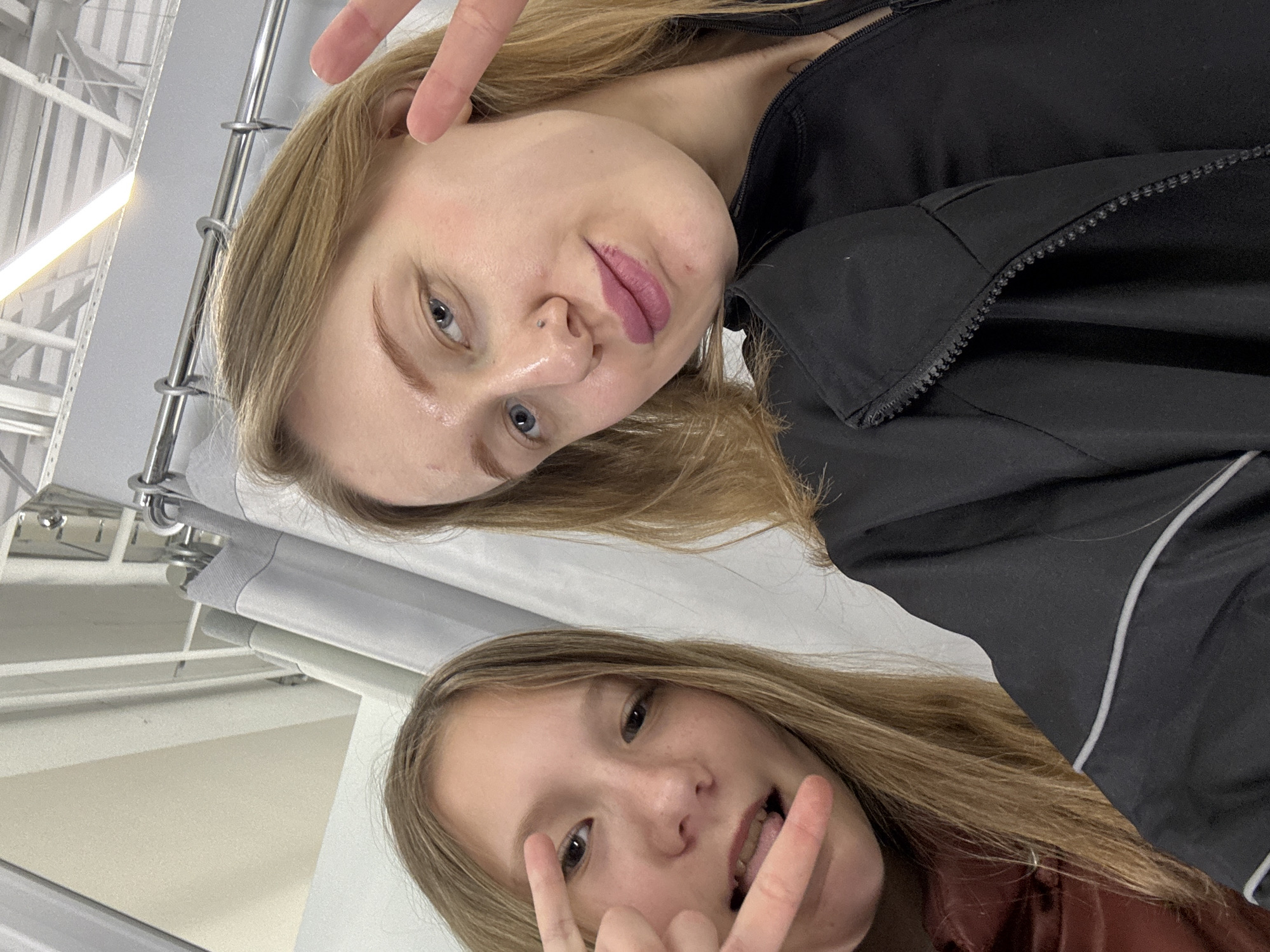 Two young women are posing for a selfie. The woman at the top of the frame has blonde hair and blue eyes. She is wearing a black jacket. The woman at the bottom of the frame has brown hair and brown eyes. She is wearing a reddish brown jacket. Both women are making hand gestures. The location appears to be indoors under fluorescent lighting, possibly in a retail or commercial space. There is no clear indication of the city of Ovoshchnoy, Russia.