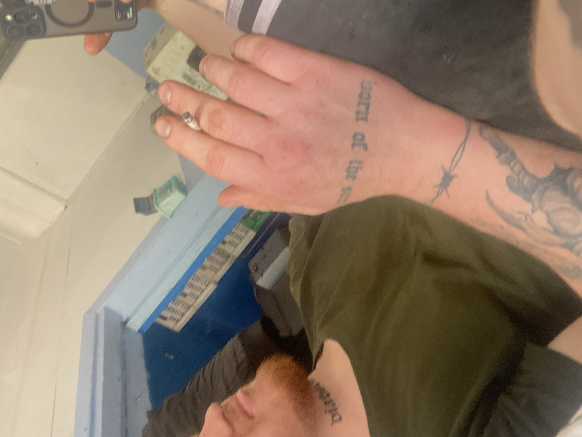 A person with red facial hair and tattoos is lying down, holding a cigarette in their right hand. A phone is visible in the upper right corner of the image. The person is wearing a green t-shirt. The background appears to be an indoor environment with blue and white surfaces, possibly a room or a structure.