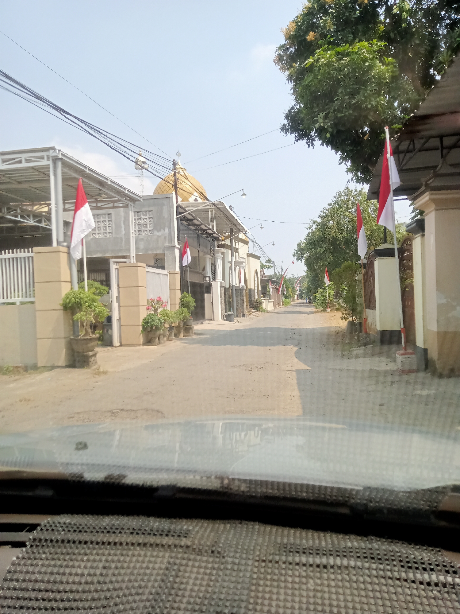 The image captures a street scene in Simo, Indonesia, under a clear sky. The perspective is from inside a vehicle, looking forward through the windshield, which is partially visible in the foreground. The street is paved with a rough, dusty surface. On both sides of the street, residential buildings and fences are visible.
Several Indonesian flags (red and white) are displayed on poles, suggesting a national holiday or celebration. Some buildings feature modern architectural elements, including metal roofs supported by columns, and ornate gates. A prominent golden dome, likely belonging to a mosque, is visible in the background, to the left of the street.
The street is lined with trees and potted plants, adding greenery to the urban environment. There are no people clearly visible in the scene, and no specific actions or interactions are depicted. The overall impression is a quiet street in a residential area of Simo, decorated with national symbols.