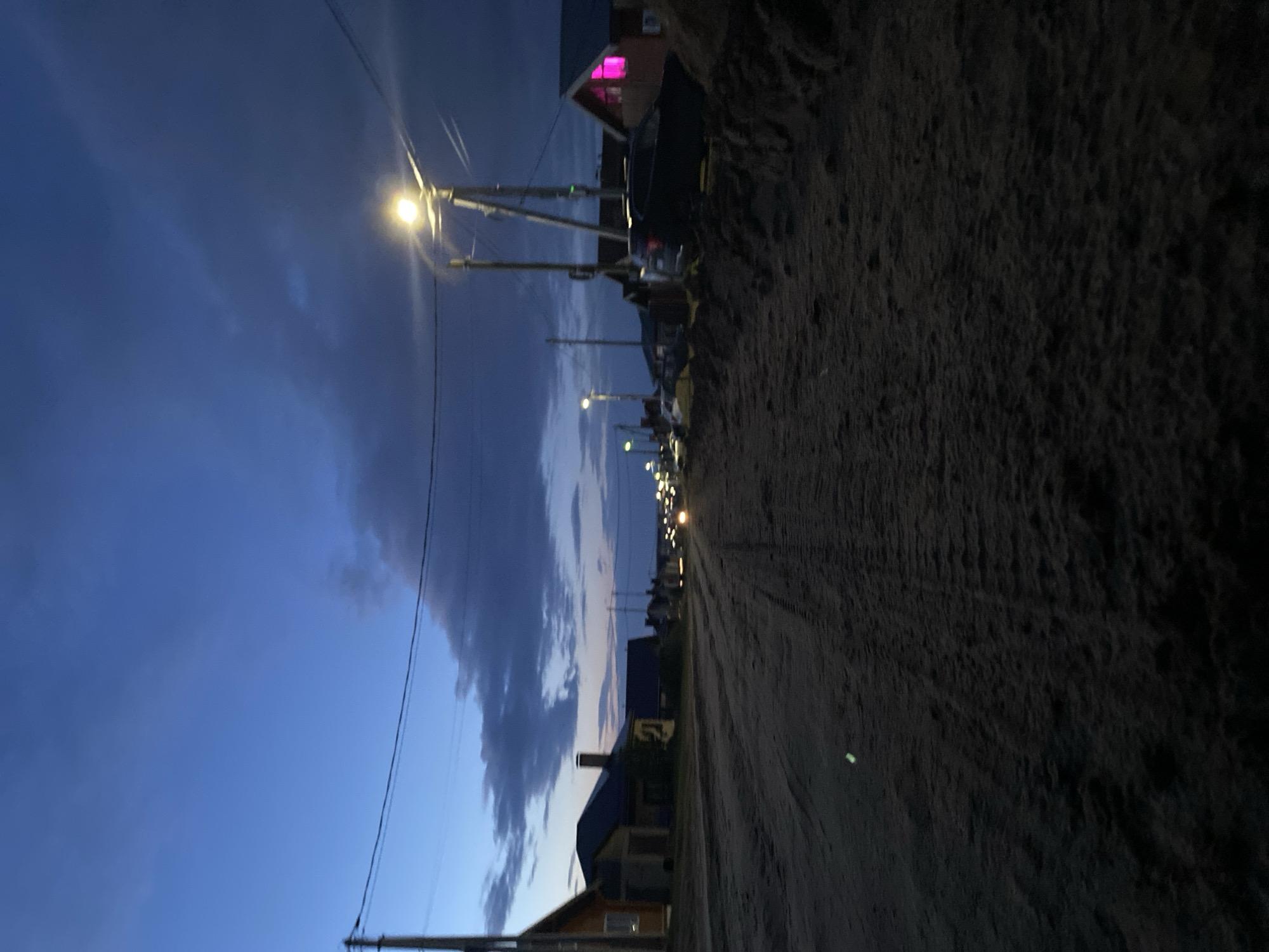 The image, likely taken in Chaikovsky, Russia, depicts a serene evening or early night scene along an unpaved road. The sky is a deep, dark blue, characteristic of twilight, with soft, scattered clouds visible above. A line of streetlights extends into the distance, brightly illuminating sections of the road and the adjacent areas with a warm glow.
Along both sides of the road, various houses are discernible against the darkening sky. One house, located on the right side further down the street, features a prominent, vibrant pink light shining from an upper window, providing a striking color contrast to the predominantly cool tones of the sky and the warm streetlights. Utility poles with overhead wires run parallel to the road, contributing to the semi-urban or rural setting.
The road itself appears to be made of rough, textured dirt or gravel, with its dark surface occupying a significant portion of the lower and right frame. There are no people or specific activities taking place, creating a quiet and still atmosphere. No visible text is present in the image.