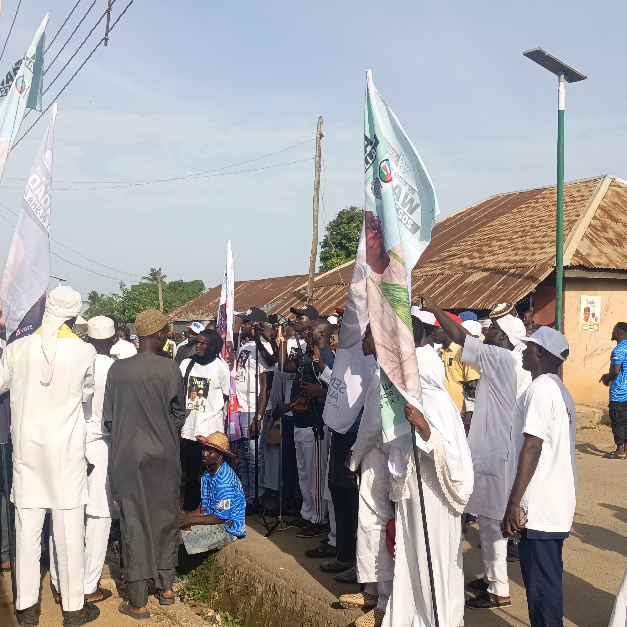 Akwanga, Nigeria, appears to be the location of this outdoor scene showing a crowd of people. Several individuals hold up political flags. A few wear white clothing and caps. A man sits on the edge of what looks like a small drainage ditch wearing a blue patterned shirt and straw hat. The background shows buildings with rusted corrugated metal roofs and a solar-powered street light. Utility lines are visible overhead.