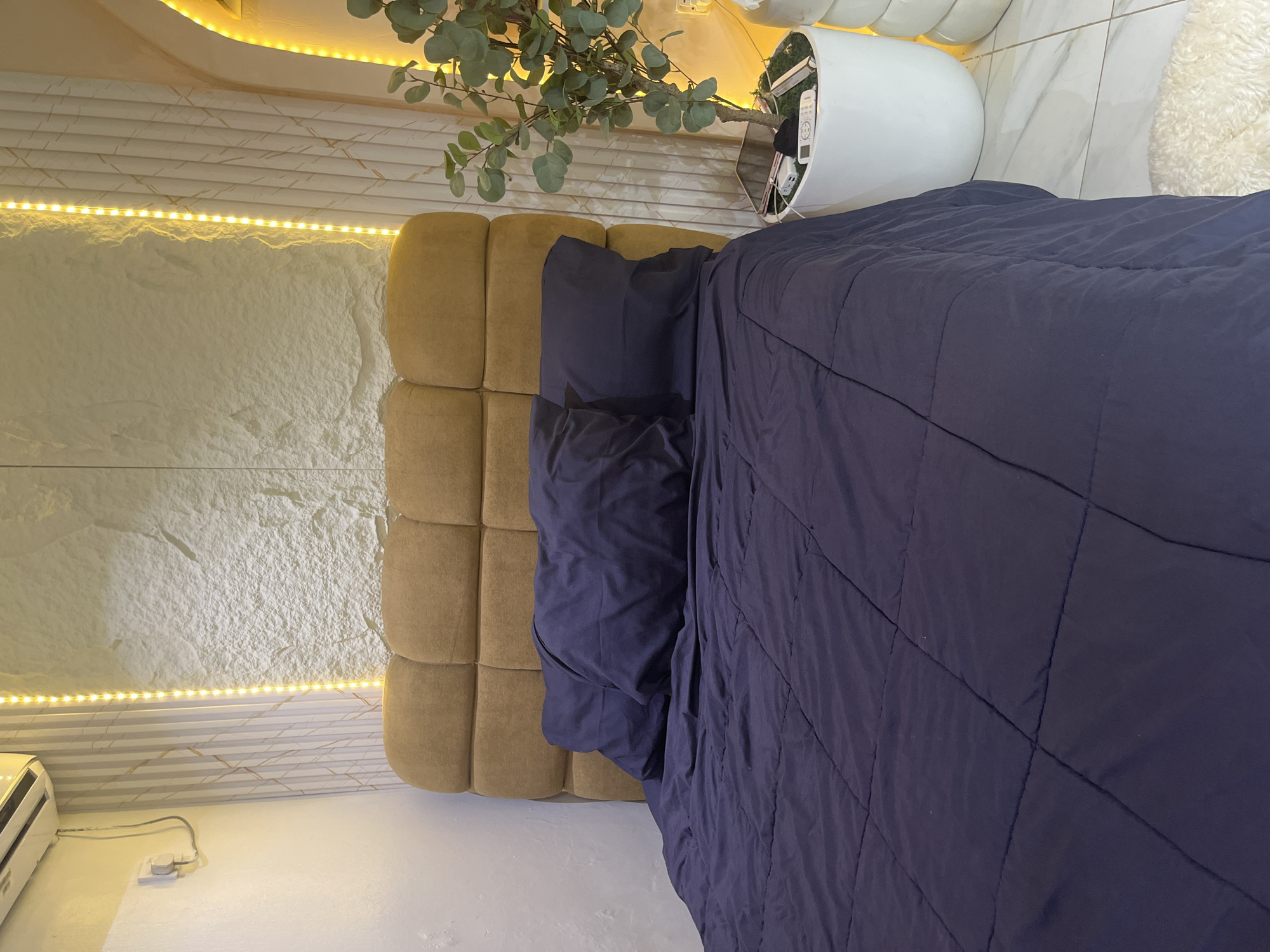 This is a medium shot, eye-level, indoor photograph taken in Abuja, Nigeria, depicting a bedroom scene. The primary subject is a bed with a dark navy comforter and two pillows. The bed has a square-tufted headboard upholstered in a mustard-yellow fabric. To the right of the bed, a tall, white planter contains a faux eucalyptus plant. The planter sits on a white surface next to a white potted plant with stacked circular tiers. Yellow LED strip lights are mounted vertically on the wall behind the headboard and also appear to be illuminating an arched doorway or alcove in the background. The wall behind the bed has a textured finish resembling rough stone. To the left of the bed, a white wall features an air conditioning unit and a power outlet with a cable. The floor is tiled with white marble.