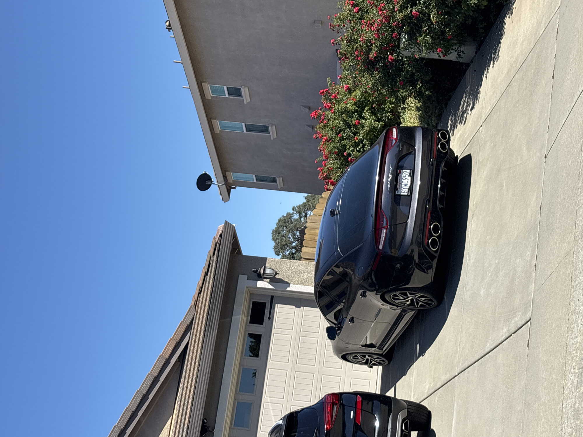 The image shows two black cars parked in a driveway in front of a house in Roseville, California, USA. The car in the foreground is a sedan with a black spoiler and quad exhaust pipes. The car behind it appears to be an SUV. The house has a beige garage door and a light gray stucco exterior. There are red rose bushes planted in front of the house, and a clear blue sky is visible above.