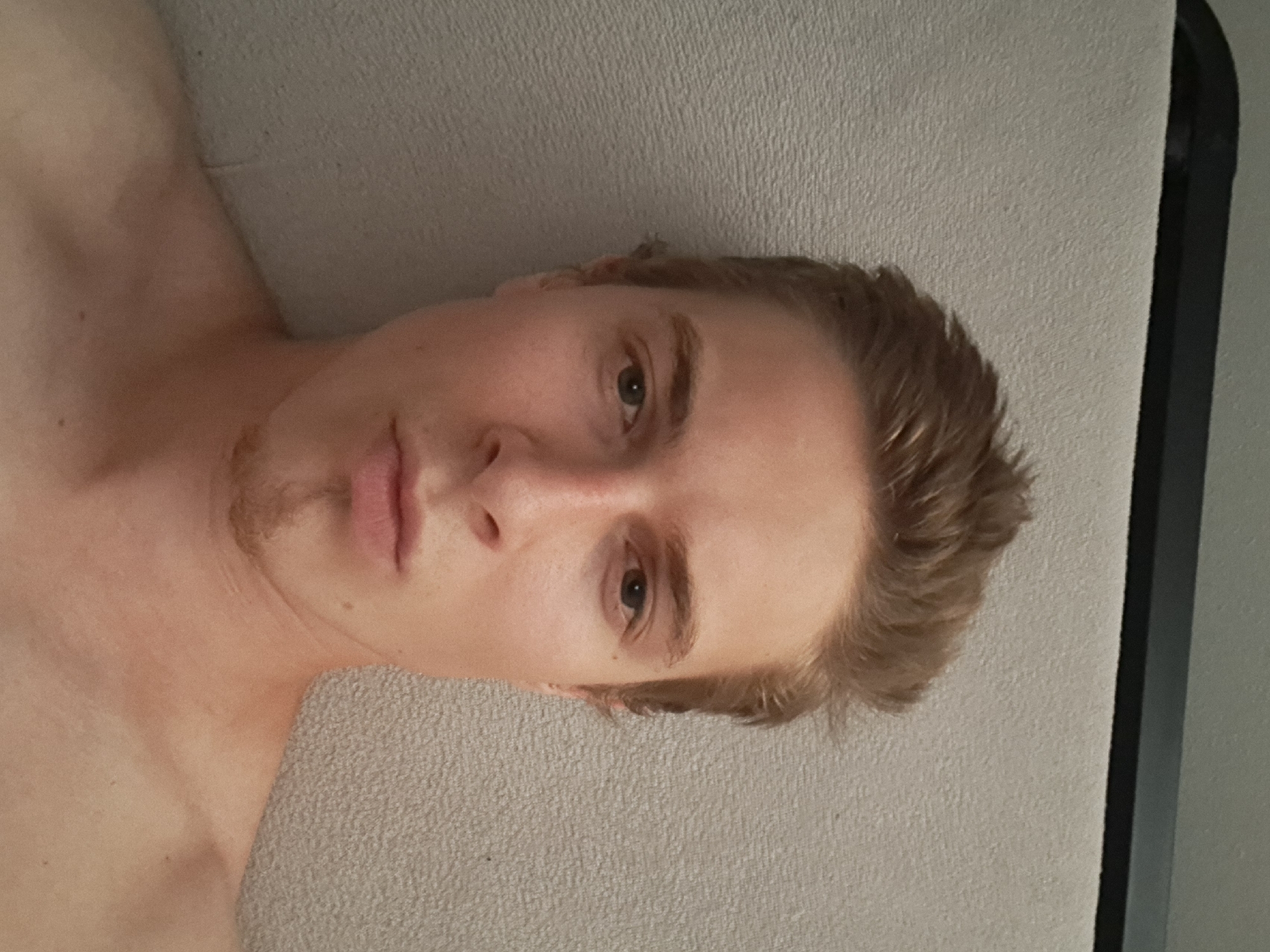 The image is a close-up, frontal selfie of a young Caucasian male with light brown hair styled upwards. He has fair skin, light brown eyebrows, and blue eyes. A faint stubble is visible on his chin. He is wearing a light-colored shirt, the top portion of which is visible around his neck and shoulders. The background consists of a textured, light gray surface. The image appears to be taken indoors. There is no identifiable information within the image to suggest a location in Chrudim, Czechia.