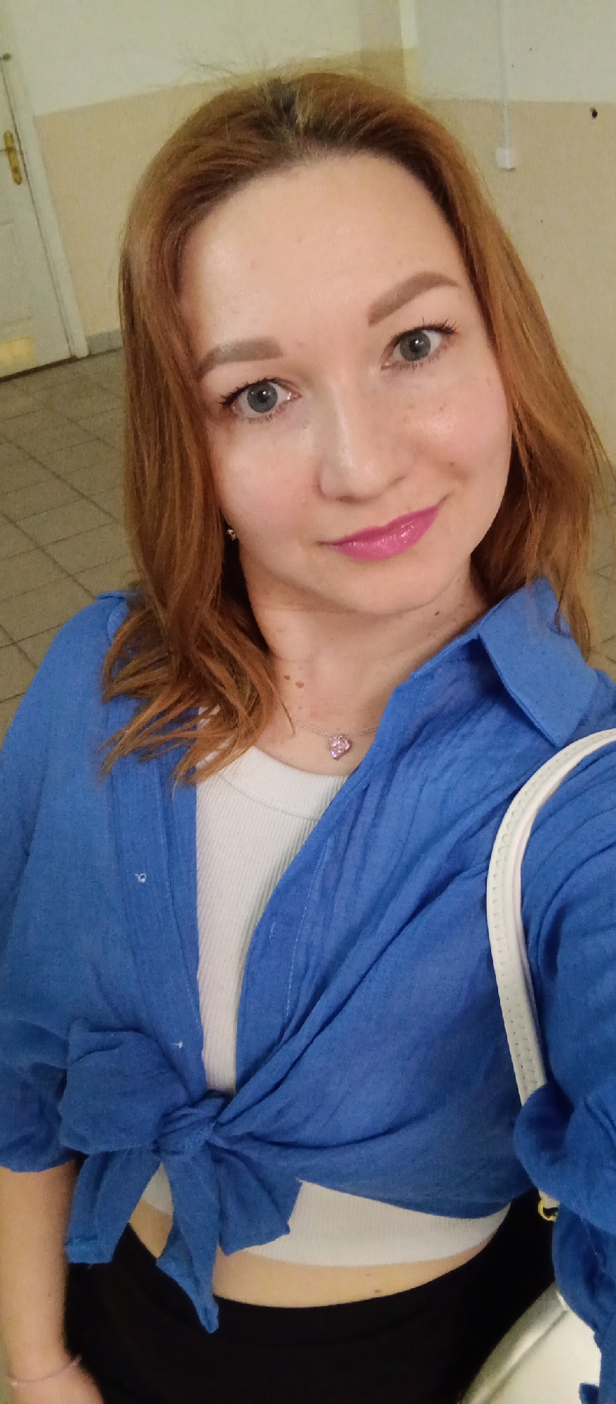 A woman is taking a selfie in what appears to be an indoor space. The lighting is moderate, casting soft shadows. She has auburn hair, fair skin with light freckling, and blue eyes. Her lips are glossy with pink lipstick. She is wearing a white tank top under a bright blue, button-down shirt that is tied at the waist. She also wears a delicate necklace with a pink pendant. A white shoulder strap suggests she is carrying a bag. In the background, the flooring appears to be tiled, and a door and wall are visible, suggesting an interior space, possibly in the city of Unknown, Unknown.