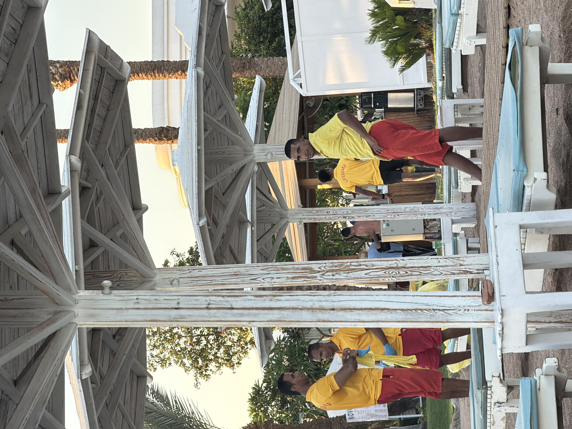 The image depicts a beach scene in Sharm El Sheikh, Egypt, with several individuals in yellow shirts and red shorts, presumably lifeguards, attending to beach chairs and umbrellas. The setting appears to be a resort, with palm trees and white resort buildings visible in the background. The lighting suggests it might be late afternoon or early morning, with a warm, soft glow.
In the foreground, two men wearing yellow polo shirts and red shorts are interacting. One man is holding a yellow cloth and appears to be cleaning or tidying up. The other man, also in a yellow shirt and red shorts, stands beside him, looking towards the camera.
Further back, another man in a yellow shirt with "LIFEGUARD" printed on the back is visible, facing away from the camera. He is standing near a series of white lounge chairs with blue cushions, which are arranged on a wooden deck. More lounge chairs and thatched umbrellas are scattered throughout the scene.
In the middle ground, several more individuals are present, including a woman in a light blue shirt and a man with his back to the camera. Behind them, a white structure, possibly a service building or part of the resort, is visible. The environment is sandy, indicative of a beach, and the overall atmosphere seems to be one of service and preparation for guests.
