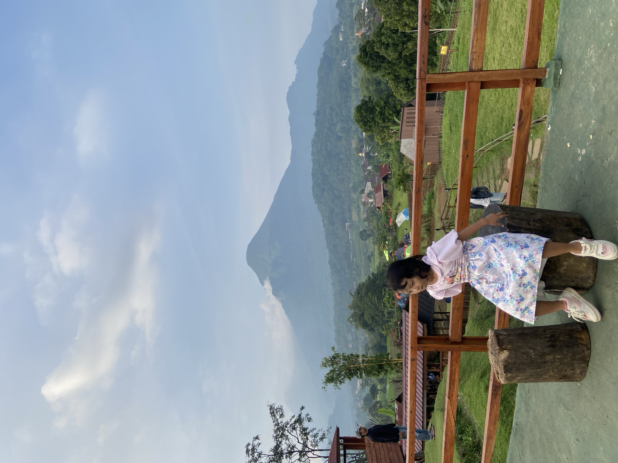 A girl wearing a light pink top and a light blue floral dress is leaning over a railing made of wooden planks and sitting on a pair of tree stumps. The railing is positioned on an elevated platform with a green surface. In the background, a mountainous landscape is visible, with trees, buildings, and tents dotted across the terrain. The sky is a light blue color with some clouds. The location appears to be in or near Tamiajeng, Indonesia.