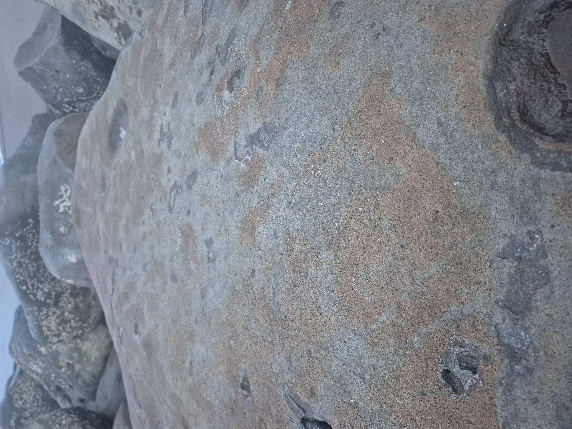 The image depicts a close-up of a large, weathered rock surface. The rock is primarily grey-brown in color, with areas of both lighter and darker shades. The surface is textured and uneven, showing signs of erosion and weathering. There are also several distinct holes or indentations visible on the rock surface. To the left side of the image, there are other rocks piled together. The information about the location in Lhokseumawe, Indonesia, is not evident from the image itself. Therefore, I can't provide a description that verifies the location based on what's visible.