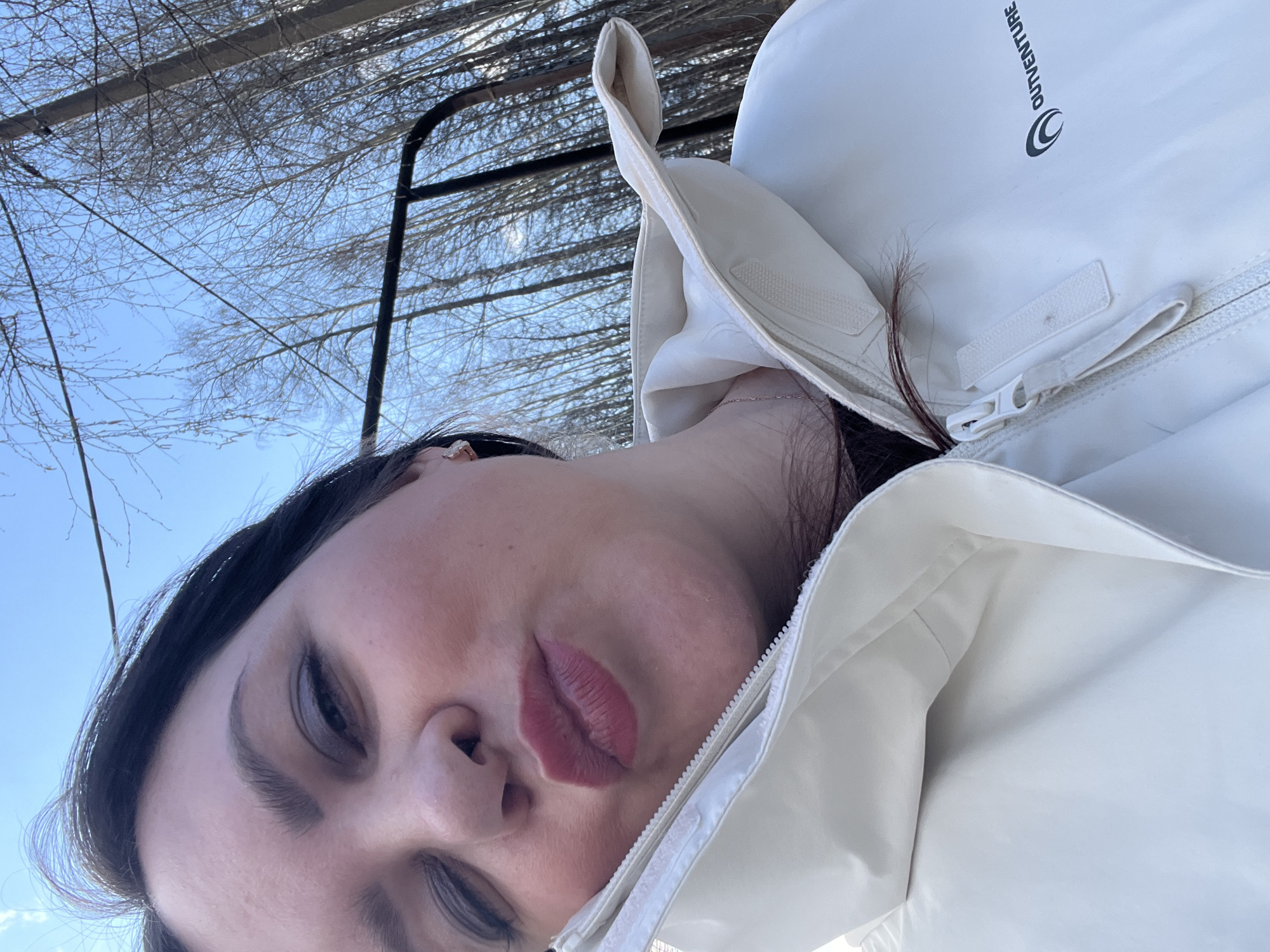 The image is a close-up selfie, rotated approximately 90 degrees clockwise, featuring a person from the chest up. The individual, likely female, has dark hair and is looking downwards towards the camera with her head tilted significantly to the right. She is wearing a white or light-colored puffer jacket, which has a visible zipper and collar. On the jacket's upper left shoulder, the brand name "OUTVENTURE" and its circular logo are clearly visible in dark text. A delicate silver chain necklace is also noticeable around her neck.
Her face shows dark, defined eyebrows and full, rosy lips that are slightly parted. The background consists of a clear, bright blue sky dotted with numerous bare tree branches, indicative of a winter or early spring season. A dark, possibly metallic, structure or frame runs vertically in the background on the right side, interspersed with the trees. The bright, natural light suggests the photo was taken during the daytime. The outdoor setting with bare trees aligns with the provided location context of Noviy, Russia. The person's expression appears neutral, perhaps a subtle pose for the selfie.