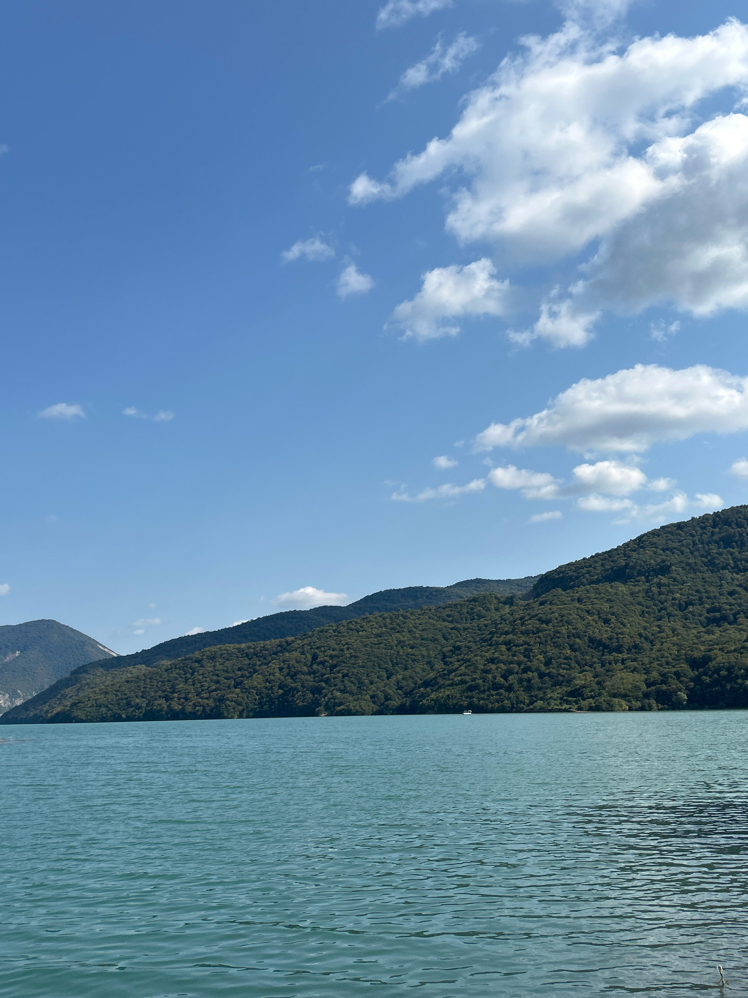 A serene, wide-angle shot captures a tranquil lake with a boat in the distance, framed by lush, tree-covered mountains under a clear blue sky dotted with fluffy white clouds. The water, a vibrant turquoise, exhibits gentle ripples that catch the sunlight, creating a shimmering effect. The mountains, cloaked in dense foliage, rise majestically from the lake's edge, their peaks partially obscured by the clear blue sky. The scene conveys a sense of peace and natural beauty, likely representing a picturesque landscape in Pkhundavi, Georgia.