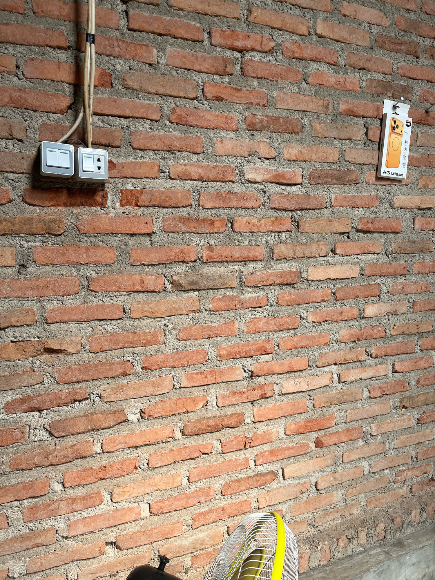 The image shows a close-up of a red brick wall, with a light gray mortar filling the gaps between the bricks. In the upper left quadrant of the image, there is an electrical outlet with two white light switches and a socket, attached to the wall by a white conduit. To the right, hanging from a nail on the wall, is a small yellow package with branding that appears to say "AG Glass" and an image of a mobile phone. In the lower portion of the image, a part of a white electric fan with a yellow trim is visible. The scene appears to be indoors or a sheltered outdoor area, given the presence of electrical fixtures. The lighting suggests it is daytime. This is a street view from Jalan Pahlawan, Kebumen, Indonesia.