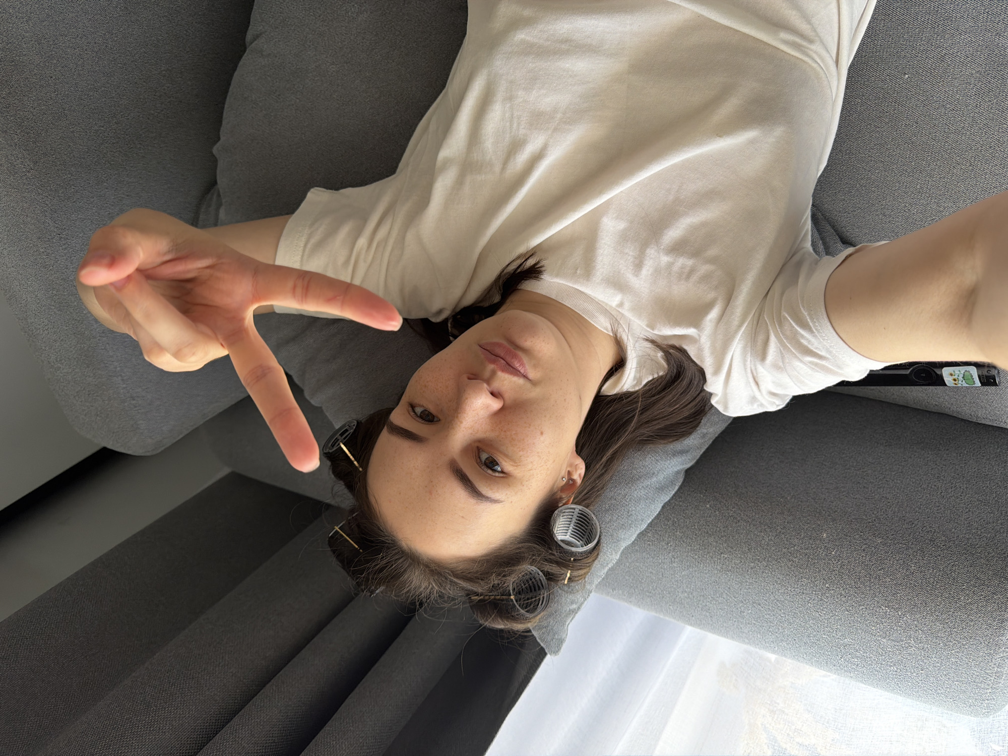 A young woman is lying on a gray upholstered couch. She is wearing a white t-shirt and has hair rollers in her brown hair. She is holding up a "V" sign with her left hand. Part of a remote control is visible on the right side of the frame. The location is not specified in the image details.