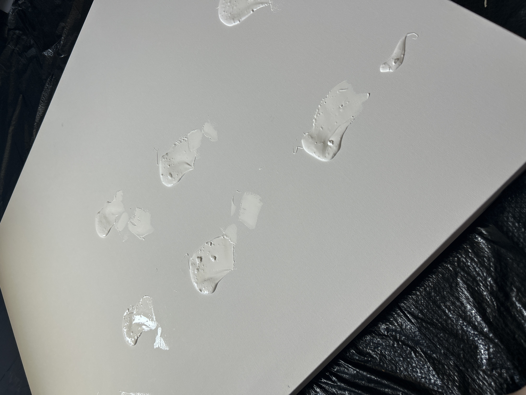 The image shows a close-up of a white canvas with several small, irregular dollops of white paint scattered across its surface. The canvas is resting on a black, crinkled plastic surface. The lighting is bright and somewhat diffused, highlighting the texture of both the canvas and the paint. There is no evidence to suggest the location of the image, and therefore Cape Coral, United States of America cannot be ascertained.