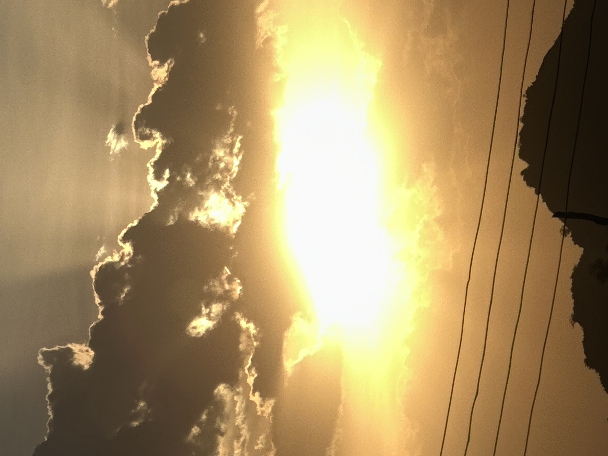 The image depicts a bright, hazy sky with the sun shining intensely through a gap in the clouds. To the right of the bright light, there are electrical wires visible, along with a dark silhouette of trees or foliage. A large cloud formation is on the left side of the image. The sky has a golden hue. It is difficult to confirm the location to be Bukuru, Nigeria based only on the image.