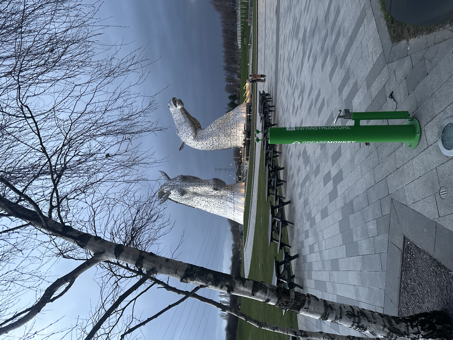 The image depicts an outdoor scene at dusk or early evening, located in Helix Park, Falkirk, United Kingdom, dominated by two large, illuminated, silvery metallic horse head sculptures known as "The Kelpies."
In the immediate foreground, to the left, is the trunk and bare branches of a birch tree. The ground consists of a paved area featuring light and dark grey rectangular pavers, upon which are arranged multiple dark-colored wooden picnic tables with attached benches, all currently unoccupied. To the right of the paved area stands a bright green cylindrical public bicycle repair station, marked "GREASEMONKEY" and "INFRASTRUCTURE" vertically, equipped with a bicycle pump and tools. A small circular ground light is visible near its base.
Behind the paved area and picnic tables, a manicured green lawn extends towards the sculptures. A curved paved pathway or road traverses this grassy expanse.
The midground is occupied by the two monumental horse head sculptures. The sculpture on the left faces primarily forward, angled slightly to the right. The sculpture on the right features a raised head with a slightly open mouth, angled towards the right. Both sculptures are illuminated from their bases with warm white light.
In the background, beyond the sculptures, the landscape includes scattered leafless trees and visible utility poles with power lines on the far left. To the far right, a small group of three indistinct individuals is observed standing on a paved area, oriented towards the sculptures. The sky above is dark and overcast, suggesting an approaching night.