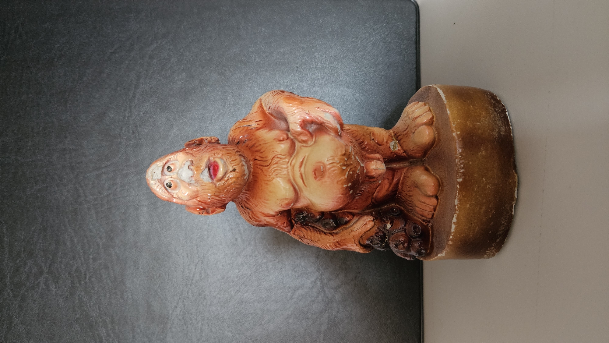 The image features an upright, reddish-orange statue depicting a corpulent, anthropomorphic figure. The figure stands on a brown, circular base, with its detailed feet and toes visible. Its body is significantly rounded, showcasing prominent rolls of flesh, especially on its large belly and chest. The arms are bent, and the hands are visible with defined fingers.
The head is round, characterized by large, dark, circular eyes. The mouth is open wide, revealing a bright red interior and white or discolored edges that suggest teeth or gums, giving the face a somewhat grotesque or highly stylized expression. The statue's color palette primarily consists of reddish-orange and brown tones, with lighter, almost creamy highlights on its more pronounced anatomical features. Darker, intricate details are present around the lower part of the figure, blending into the base.
The statue shows signs of age or wear, including minor chipping or discoloration, particularly around the facial features and along the edges of the base. It is placed on a dark, textured surface, possibly leather, which occupies the left side of the frame. A lighter, plain background, likely a wall or another surface, is visible on the right. There is no discernible activity taking place, as the subject is a static object. No text is visible in the image.