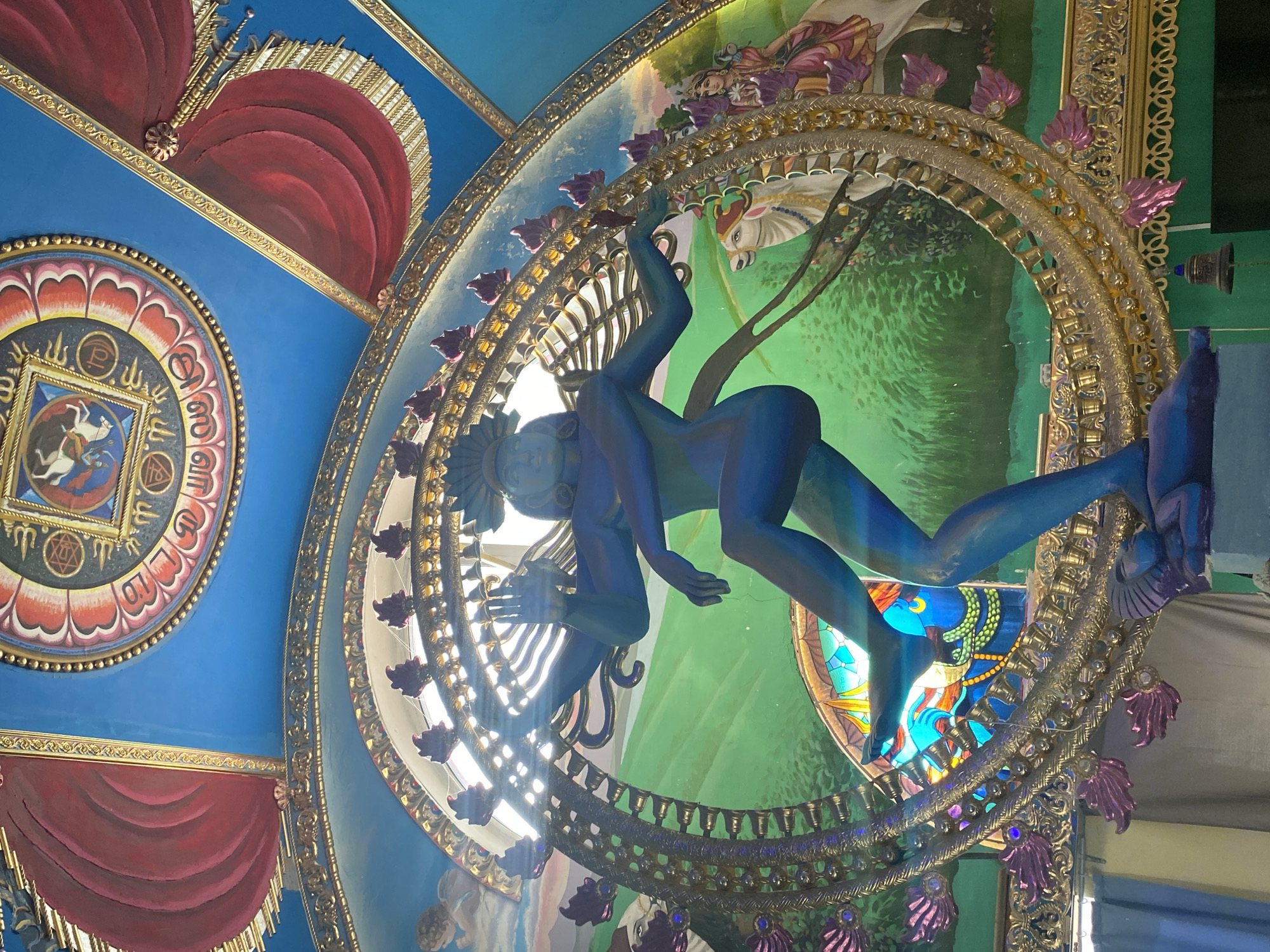 The image depicts a statue of Shiva in a dancing pose, known as Nataraja, adorned with multiple arms. The statue is predominantly blue and is set against a vibrant mural. The mural features a lush green landscape with a person riding a white bull and another figure, possibly a deity, seated. Above the central statue, the ceiling is elaborately decorated with blue paint, red drapery, and ornate golden borders. There are also circular motifs with what appear to be religious symbols and imagery. The overall scene suggests a decorative or artistic installation, likely within a temple or cultural space. The question asks for the location in Kazan, Russia, but the image itself does not contain any visual cues or text that would indicate this specific geographical location.