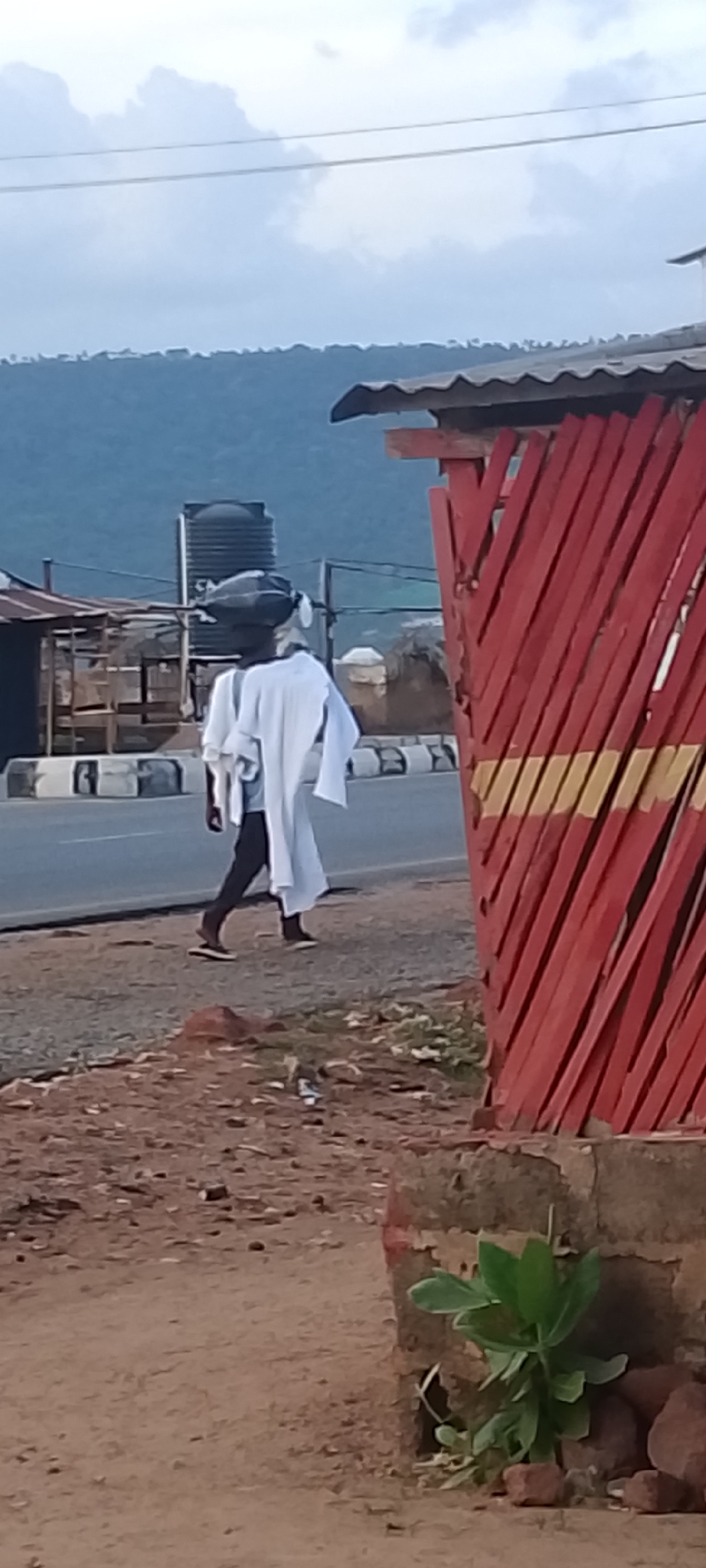In Adankolo, Nigeria, a person walks along a road carrying a dark object on their head and white cloth draped over their body. The road is bordered by a painted barrier of red and yellow stripes. In the background, there is a water tank and a landscape of hills under a cloudy sky.