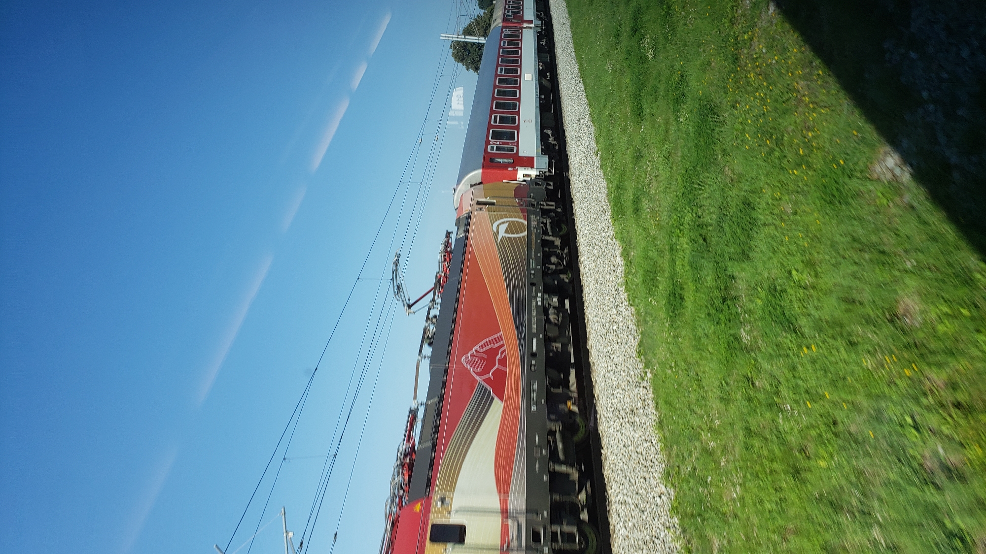 This is a side view, captured from a moving train, of a red and white passenger train traveling on a track. The train has a distinctive red locomotive with a stylized white and orange design that includes a depiction of mountains. It is connected to several red and white passenger cars. The train is traveling through a rural landscape with a clear blue sky above. In the foreground, there is a strip of green grass and a gravel track bed. The image appears to be taken on a sunny day. The location is stated to be the city of Varín, Slovakia.