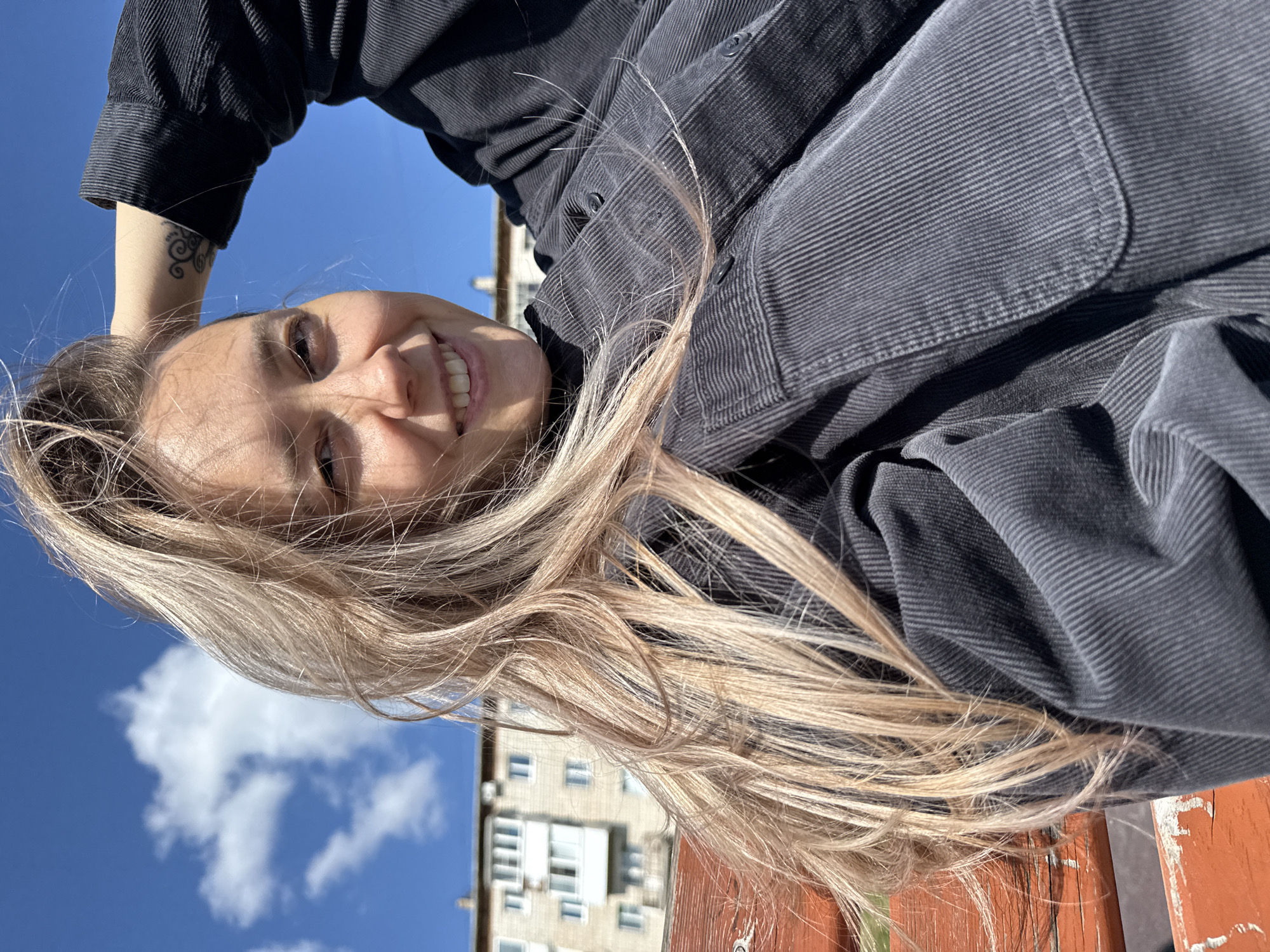 A young woman with long blonde hair is captured in a selfie-style photo. She is sitting on an orange wooden bench, with her left arm raised behind her head, touching her hair. She is wearing a dark grey corduroy shirt. She is smiling directly at the camera. In the background, there is a light blue sky with some white clouds, and a multi-story apartment building. The overall setting appears to be an urban outdoor environment, likely a park or residential area, in Gornozavodsk, Russia. The image is taken in daylight.