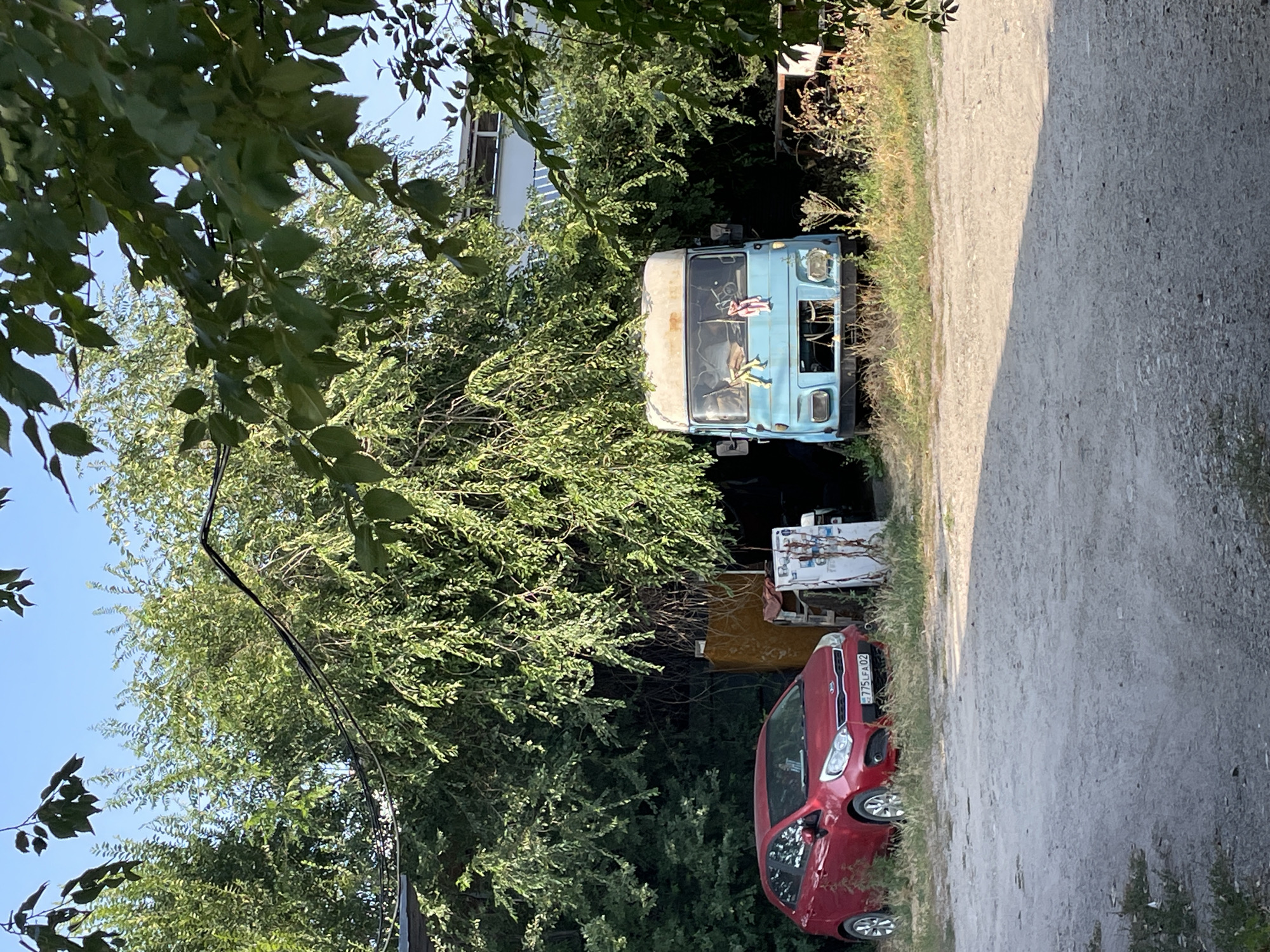 In Almaty, Kazakhstan, a red Kia Rio hatchback is parked on a gravel surface next to overgrown vegetation. To its right, partially obscured by trees and shrubbery, sits a light blue van with a white roof. The van appears old and possibly in disrepair, with a damaged front grille. A white object, possibly a refrigerator or cabinet, stands between the car and the van. The scene is bathed in sunlight, casting shadows from the trees. The sky is clear and blue.
