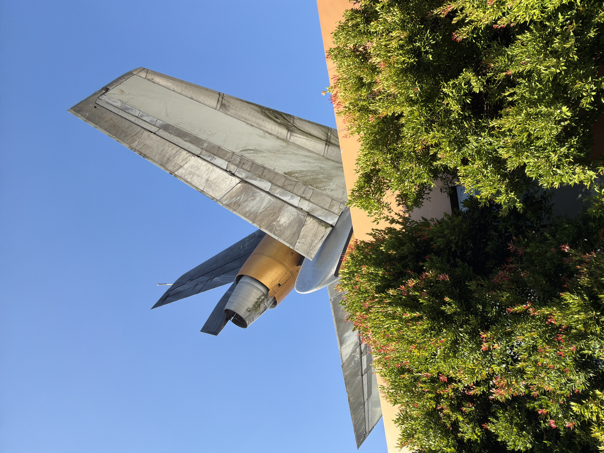 An upward shot shows the tail section of an airplane, including its engines and wings, protruding from the top of an orange building. Lush green bushes with red-tinged leaves are in the foreground, partially obscuring the building and the lower portions of the airplane. The sky is clear blue. The airplane appears to be mounted as a decorative feature on the building. The location is the city of North Kuta, Indonesia.