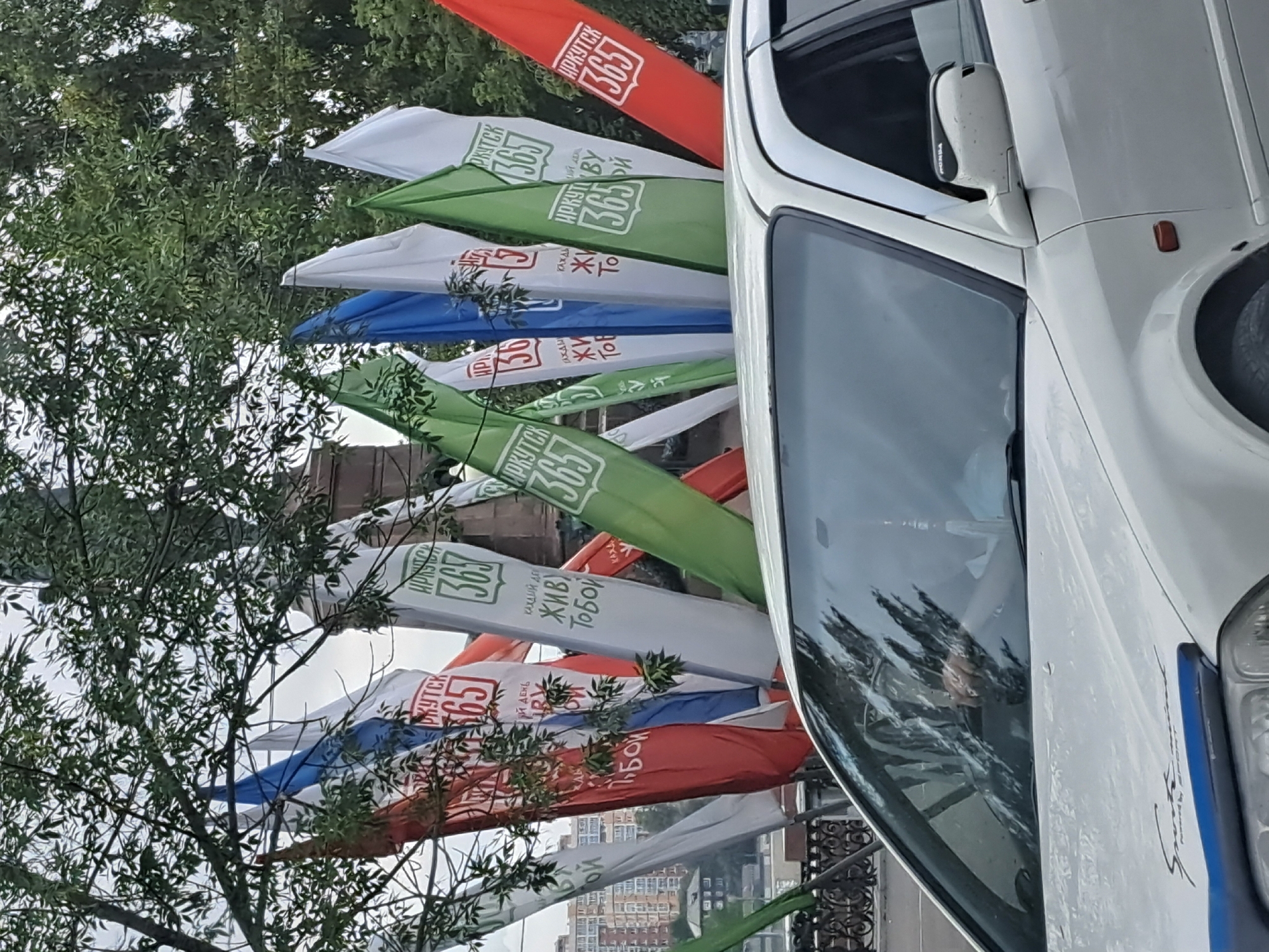 In Irkutsk, Russia, a white sedan is parked near a sidewalk adorned with numerous vertical flags. The flags, predominantly red, white, green, and blue, bear inscriptions in Russian, with visible text like "ИРКУТСК 365" and "КАЖДЫЙ ДЕНЬ ЖИВУ ТОБОЙ". Some flags also feature the Russian tricolor. In the background, a multi-story building is visible, along with trees and a pedestrian fence. The car's windshield reflects trees, and the driver, partially obscured, appears to be holding the steering wheel. The overall scene suggests a streetscape event or celebration in the city.