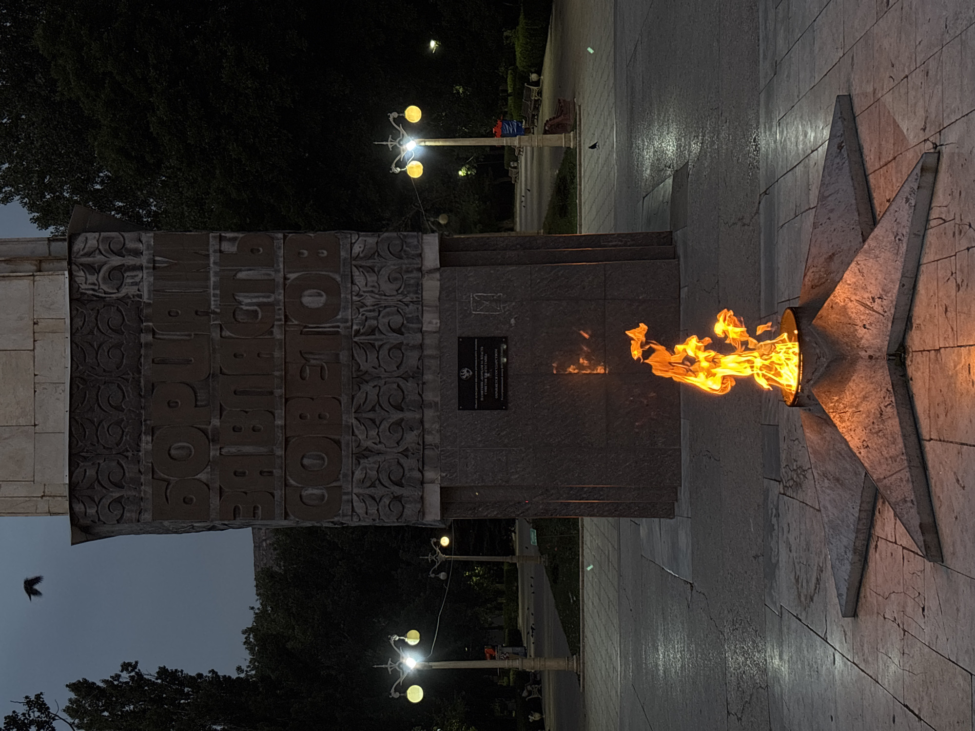 The image depicts the Eternal Flame in Makhachkala, Russia, located at the monument "To the Heroes of the Civil War and the Great Patriotic War." In the foreground, a bright orange flame burns in a star-shaped brazier, casting a warm glow on the surrounding stone paving. The brazier is set within a large, five-pointed star. Behind the flame, a tall, stone monument dominates the scene, adorned with carved text in Cyrillic script. The monument is illuminated by streetlights, one of which is visible on the left side of the image with two yellow lamps. In the background, trees create a dark silhouette against the twilight sky. The ground is paved with large, grey stone slabs, some of which are wet and reflect the light. Several small birds are visible on the ground and in the sky, adding a touch of natural life to the solemn setting. The overall atmosphere is one of remembrance and respect.