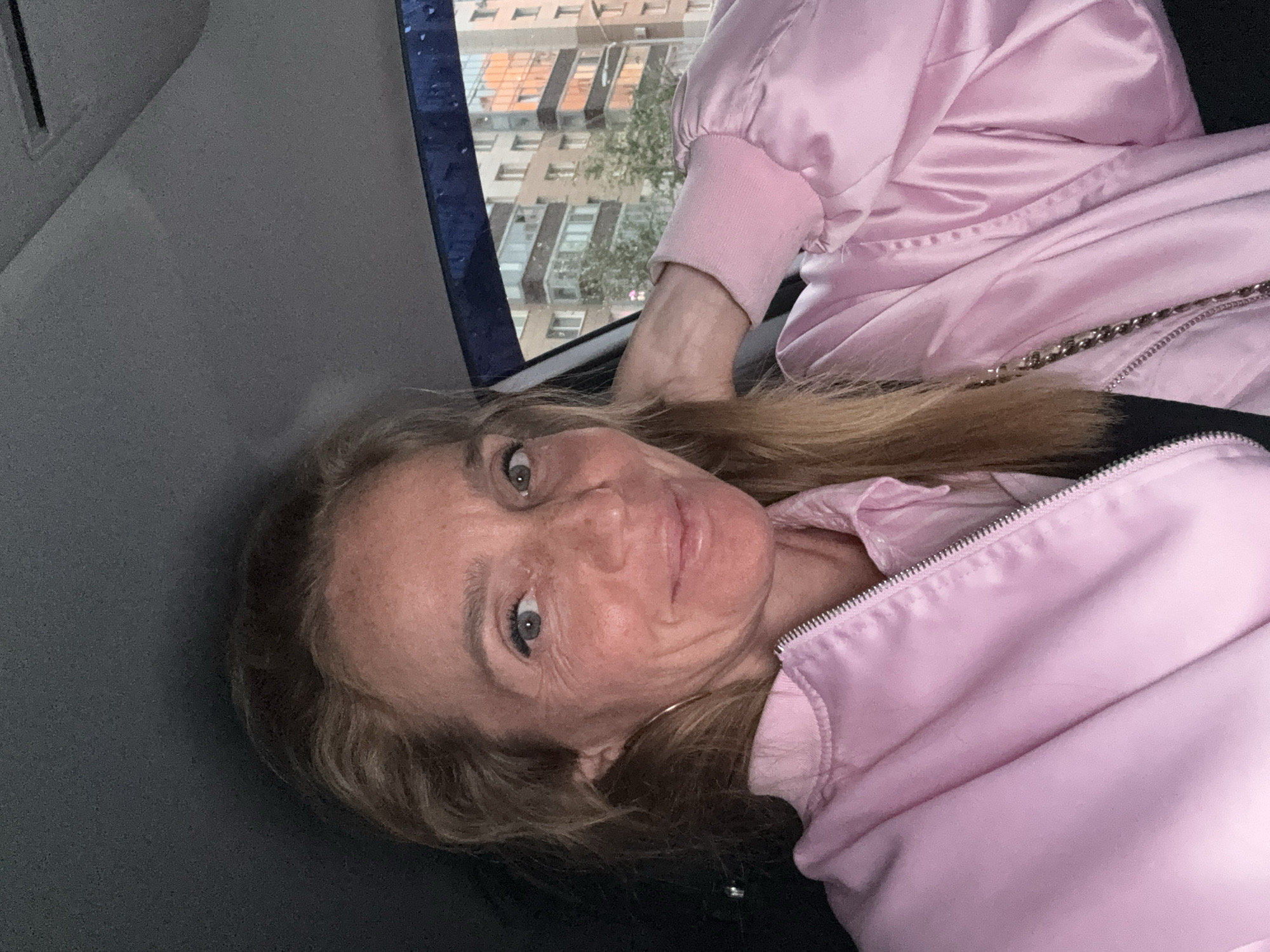 An overhead, medium shot shows a woman with blonde hair and fair skin, with freckles visible on her face. She is wearing a light pink satin bomber jacket and a dark-colored top. Her head is tilted to the left, and her right hand is resting on the back of her neck. She is looking directly at the camera with a slight smile. The background shows the interior of a vehicle, with the ceiling of the car visible above her head. Through the car window, a city street is visible with apartment buildings. The time of day appears to be late afternoon or early evening, as the light is warm and reflects off the windows of the buildings. The scene is likely in a residential area of a city, possibly Russia, given the architectural style of the buildings.