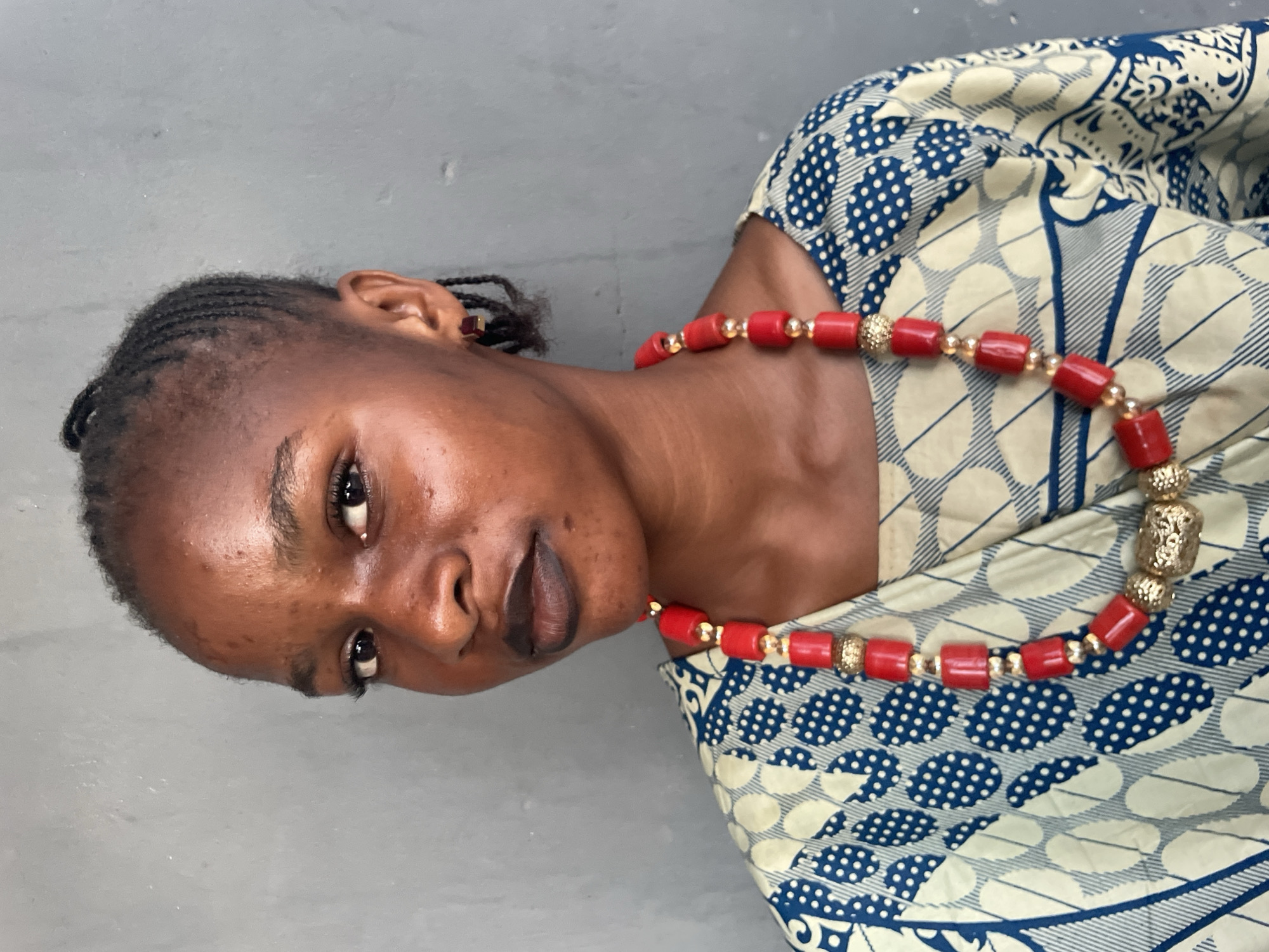 A medium shot depicts a woman with dark skin, dressed in a patterned garment of cream and blue, and adorned with a red and gold beaded necklace. The woman has braided hair. The setting is nondescript, featuring a gray wall as the backdrop. There is no visual information to connect this scene to Badagry, Nigeria.