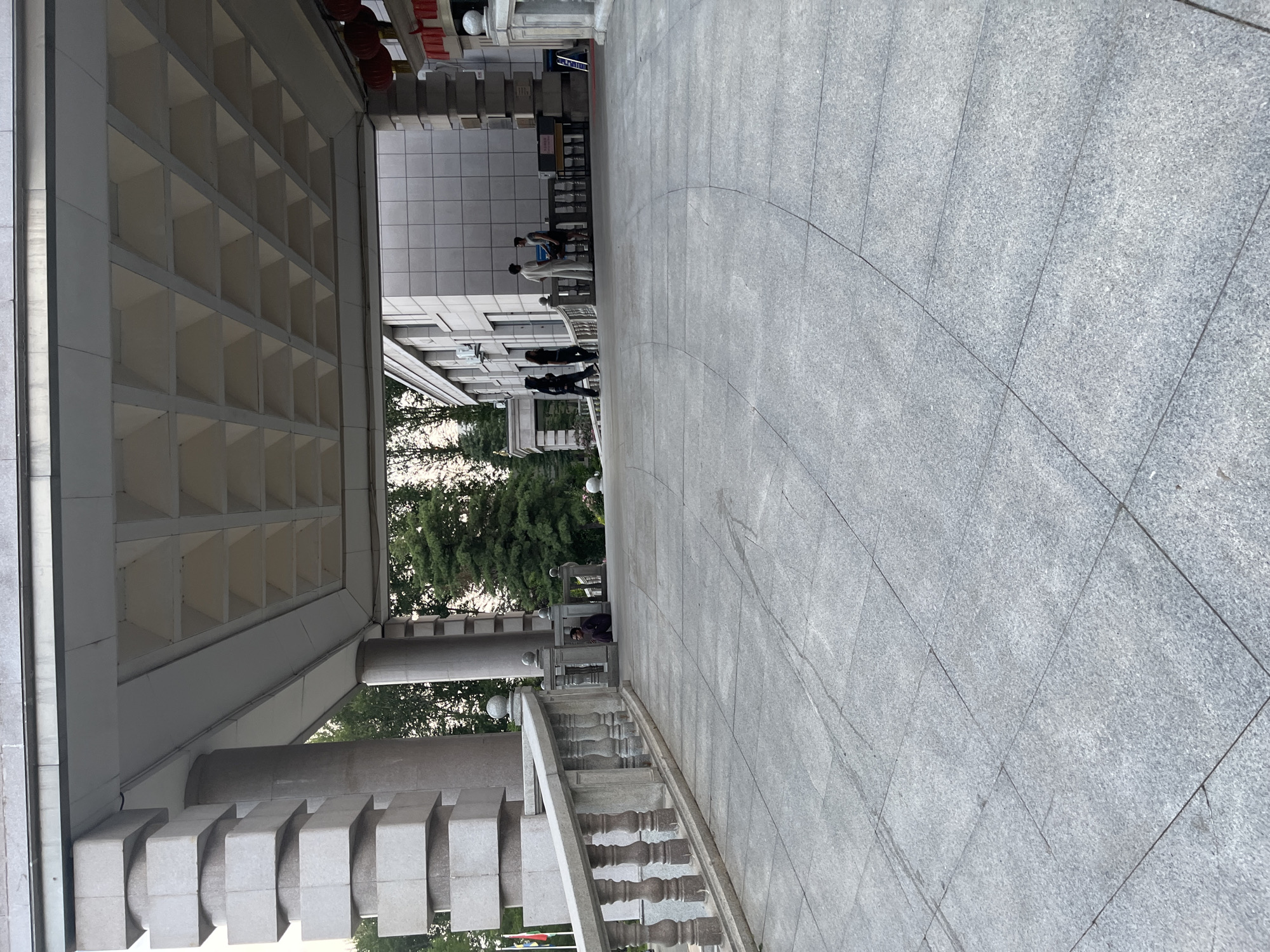 This is an eye-level, medium shot of a wide, paved walkway leading towards a building with an overhang supported by large columns. The walkway is made of grey square tiles. To the left, a stone balustrade with ornate spindles runs alongside the walkway, curving away from the viewer. Behind the balustrade, there are flags and trees.
Under the overhang, several people are visible. Three individuals in dark clothing are walking away from the viewer. Further into the scene, two people are standing and talking, with another person sitting to the far left, near the balustrade. The building itself is clad in light-colored square tiles. The overhang's ceiling has a repeating geometric pattern. Red lanterns are visible hanging from the right side of the overhang.
The scene appears to be an entrance to a building, possibly a government building or a cultural institution, given the flags and the formal architecture. The lighting suggests it is late afternoon or early evening. The presence of people suggests a public space. The architecture indicates a location in China, with the specific detail of the building resembling the Jilin Provincial Museum in Changchun City, China, based on its distinctive architectural features and style.