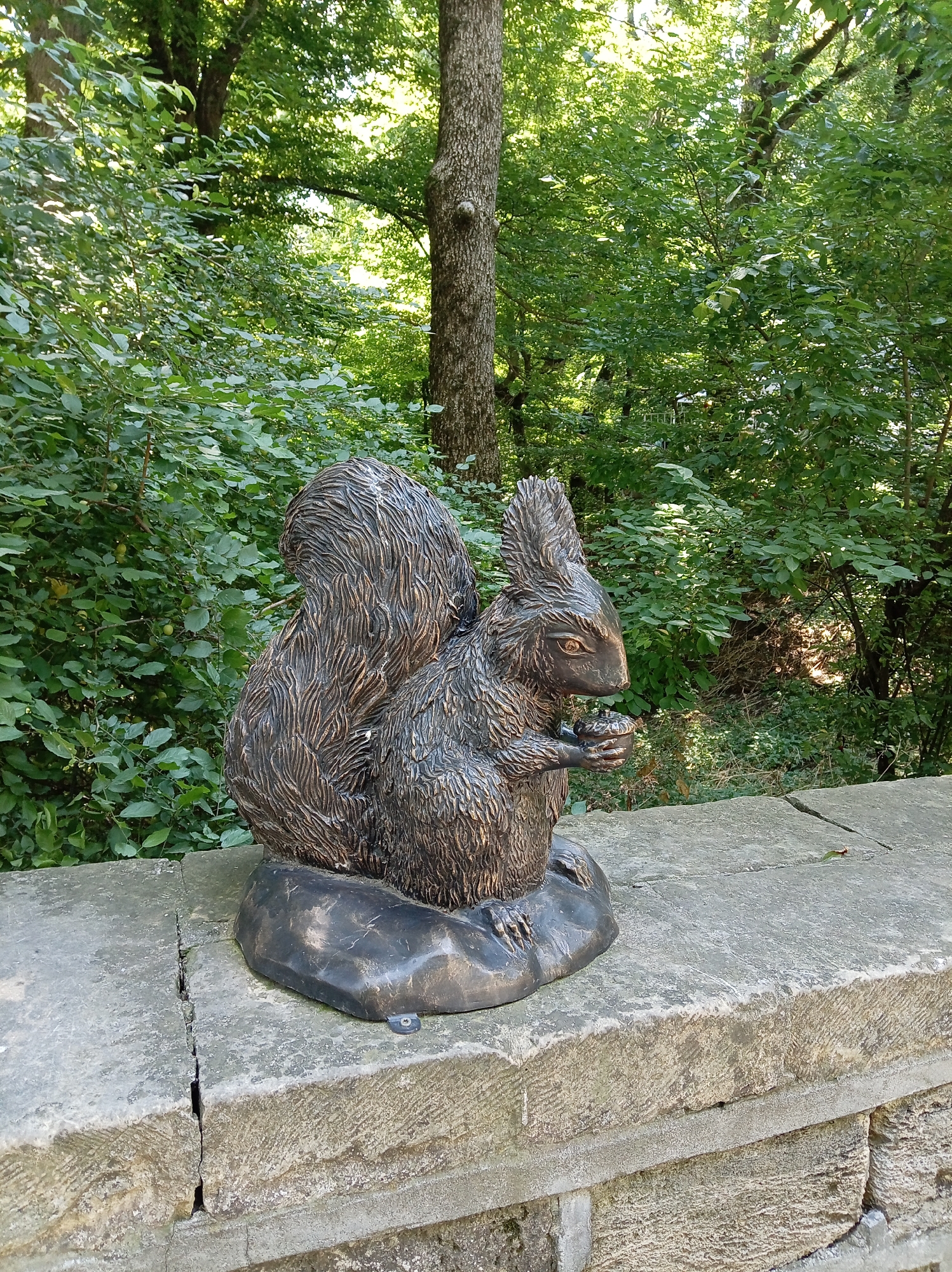 An outdoor, eye-level, full shot features a bronze statue of a squirrel holding a nut. The squirrel is dark brown with intricate fur detailing and is positioned on a dark, irregular base that sits on a cracked stone ledge. The ledge is made of weathered, light-colored stone blocks. Behind the squirrel and ledge, a dense, green forest with tall trees and lush foliage is visible, suggesting a natural, park-like setting. The lighting indicates daytime, with sunlight dappling through the leaves. There are no people or animals present in the image. This statue is located in the city of Zheleznovodsk, Russia, likely in a park or public garden.