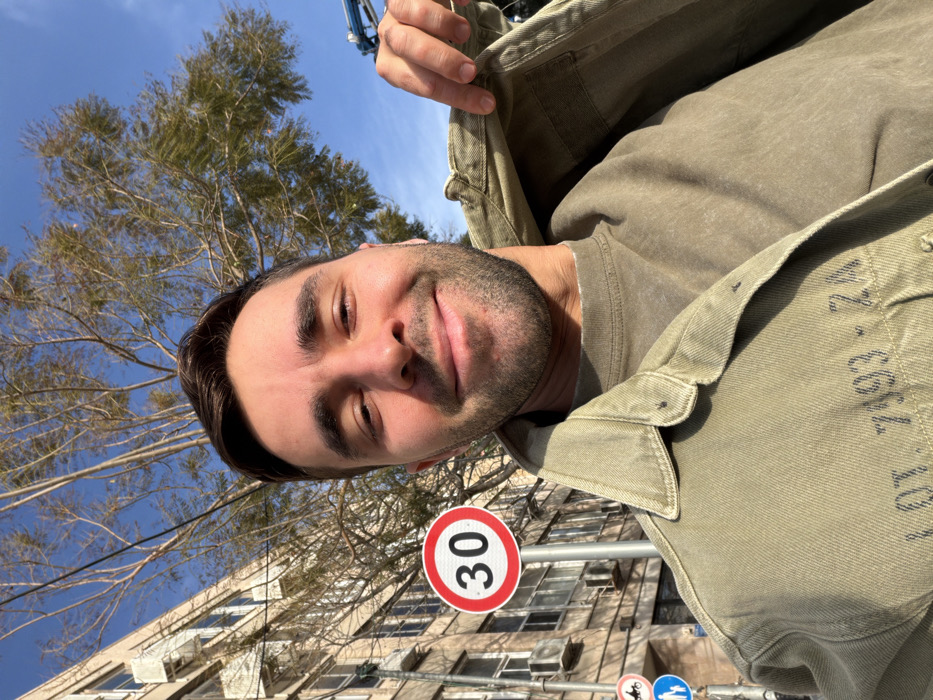 The image displays a male individual in a selfie-style shot outdoors on a clear, sunny day in an urban area consistent with Tel-Aviv, Israel. The subject, a man with short dark hair and light facial stubble, is positioned centrally, smiling directly at the camera. He wears a light olive-green crew-neck shirt and an open olive-green denim-style jacket, the lower left front of which displays the faded text "LOT. 7393 - 2A". His right hand is visible, pulling up the right lapel of his jacket.
To the left of the subject, a multi-story building with a light-colored facade, numerous windows, and multiple external air conditioning units is visible. Immediately adjacent to the building, a pole supports several circular street signs: a prominent white sign with a red border indicating a "30" speed limit, a red-bordered white sign with a black motorcycle symbol crossed by a red diagonal line (prohibiting motorcycles), and a blue sign with white pedestrian and bicycle symbols (indicating a shared path). Overhead, numerous tree branches with sparse foliage are silhouetted against a bright blue sky. In the upper right portion of the image, a section of blue and grey heavy machinery, appearing to be a hydraulic arm or boom, is partially visible. The scene is brightly illuminated by direct sunlight.
