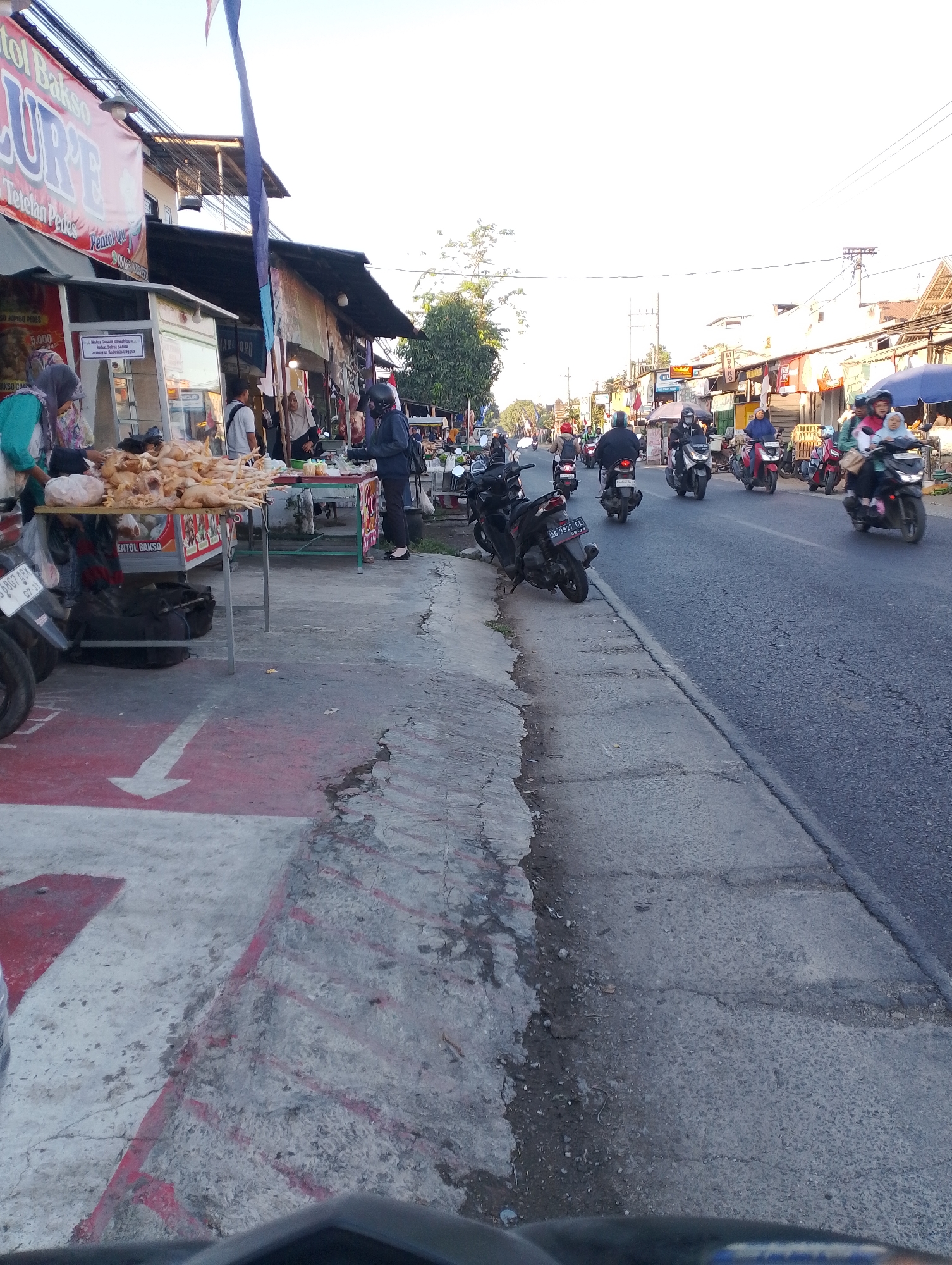 The image captures a street scene in the city of Purworejo, Indonesia. The foreground features a sidewalk with a red and white arrow painted on it, indicating direction. To the left, a food stall displays raw chickens for sale, with people interacting at the stall. Behind the stall, the facade of a building with a red banner that reads "Bakso LURE" is visible. The street itself is busy with motorcycle traffic, with several riders and passengers visible. Buildings line the street on both sides, some appearing to be shops. The time of day seems to be late afternoon or early evening, with the sun casting long shadows. The overall atmosphere suggests a typical day in a bustling Indonesian market or commercial area.