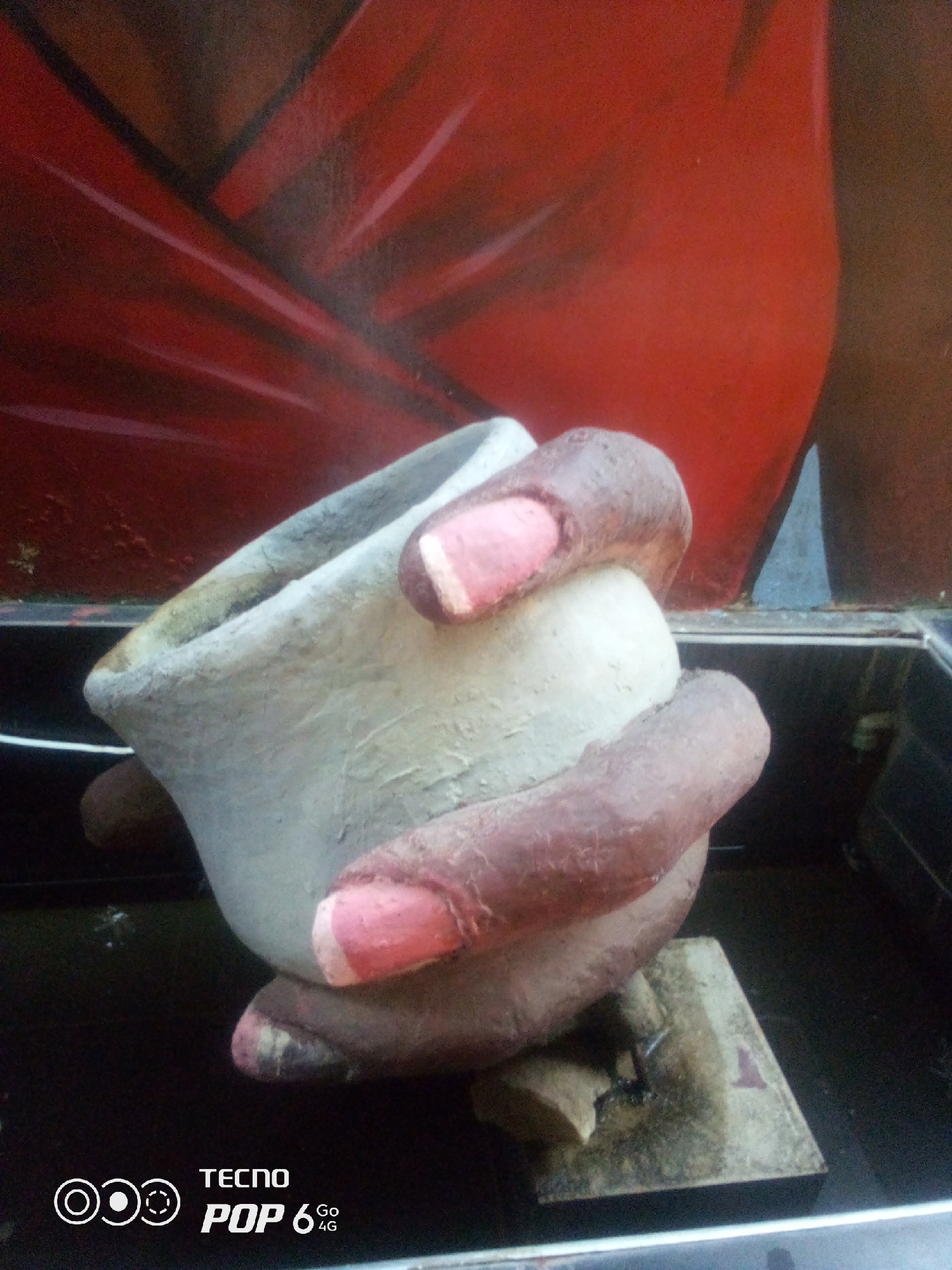 A close-up shot shows a person's hand, with dark skin and pink painted fingernails, holding a small, off-white, textured pot. The pot appears to be made of clay or a similar material and has a slightly irregular shape. The hand grips the pot firmly, with the fingers wrapping around its sides. The background is a blurry mix of red and brown, possibly a painted mural or fabric. The lower portion of the image shows a dark surface, likely a table or shelf, with a metallic edge. In the bottom left corner, a white logo and text read "TECNO POP 6 4G." The image lacks any discernible details that would indicate its location within the city of Howayi, Nigeria, or any other specific geographic area.
