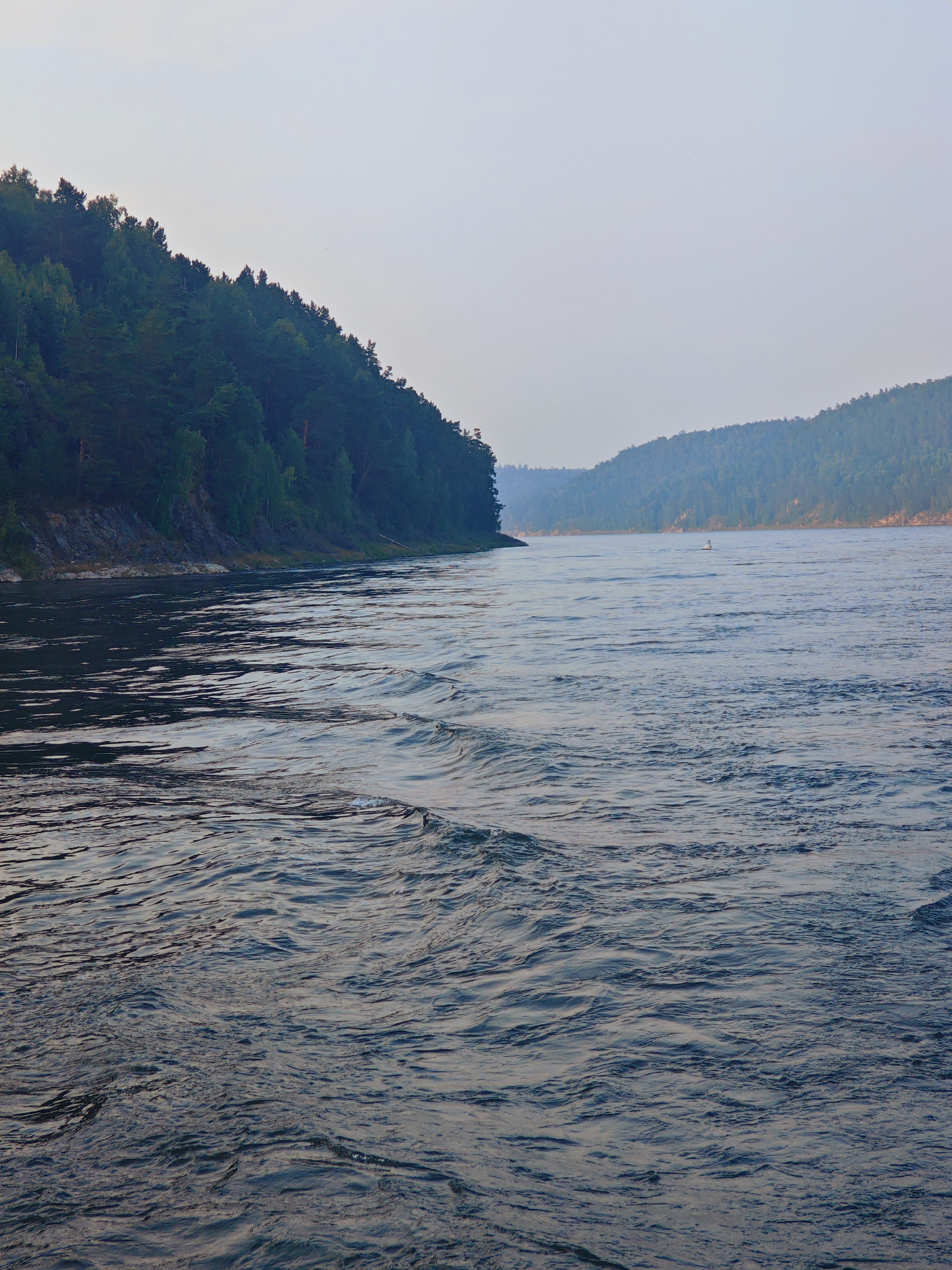 The image depicts a wide river flowing through a forested valley, likely in Predivinsk, Russia. The foreground is dominated by the dark, rippling water of the river, with visible waves creating a textured surface. On the left bank, a steep, densely forested hillside rises, with dark green evergreen trees covering its slope and rocky outcrops visible near the water's edge. The right bank of the river recedes into the distance, also lined with rolling, tree-covered hills that fade into a hazy, muted blue in the background. The sky is overcast and pale, with a soft, diffused light. In the middle distance of the river, a small, white navigational buoy can be seen. There are no people or animals visible in the frame. The overall atmosphere is one of natural serenity and vastness.