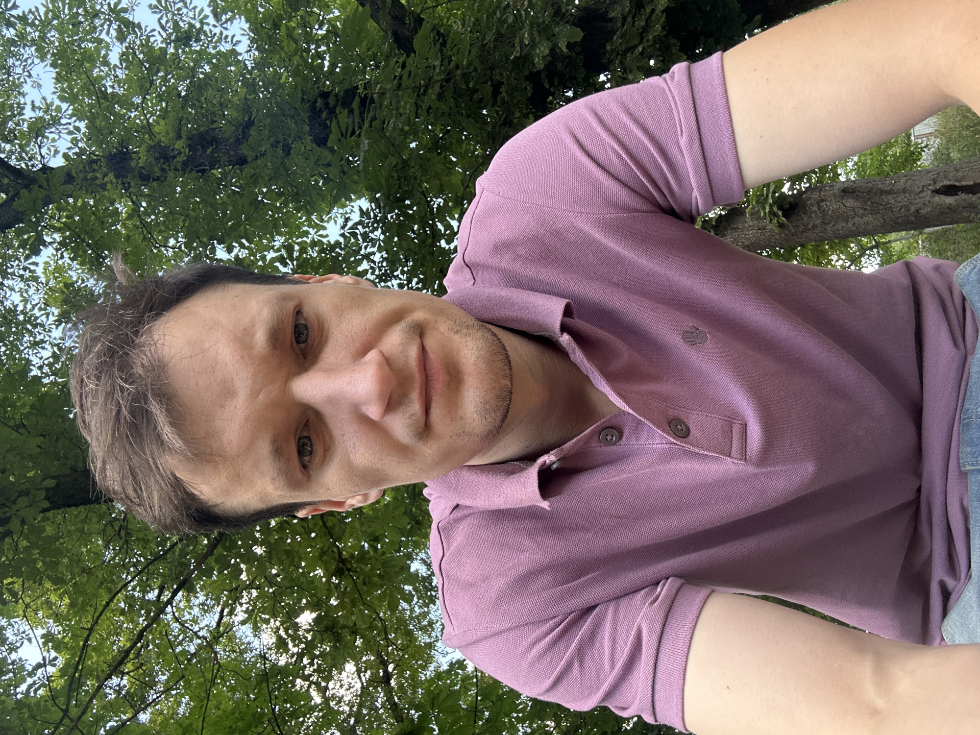 A young man with brown hair is looking at the camera, with trees and leaves behind him. He is wearing a pink polo shirt with two buttons and a small embroidered symbol of a hand on the left side of his chest. The man is outdoors, with dappled sunlight suggesting it is daytime. The location is identified as Makiv, Ukraine.