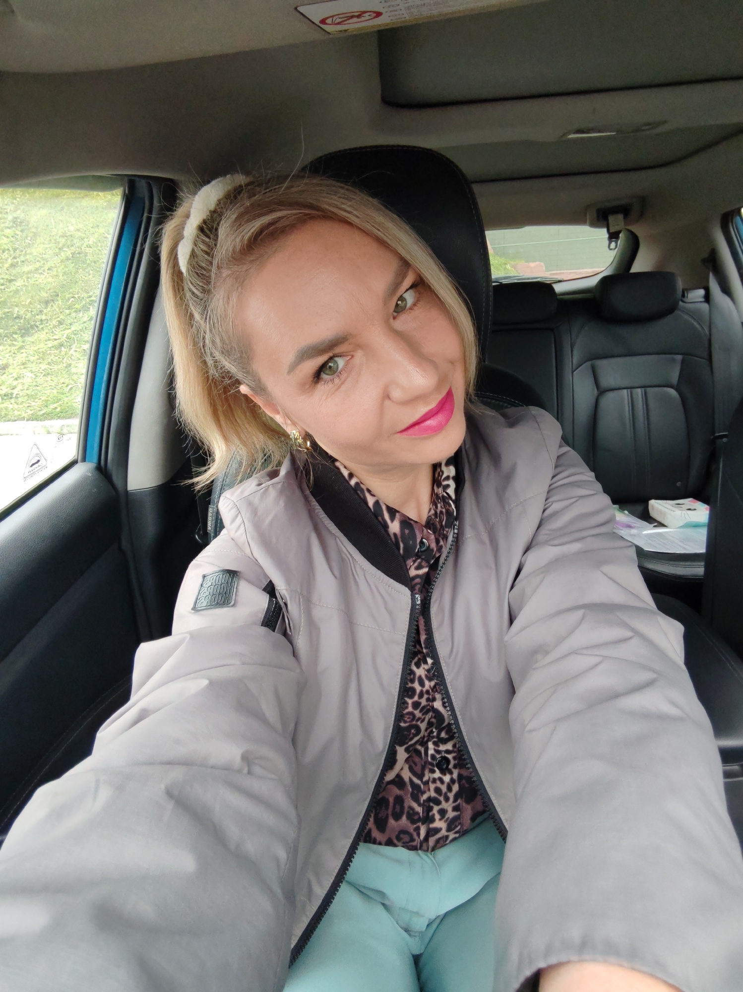 A woman with blonde hair pulled back in a ponytail, wearing a grey puffer jacket with a black and leopard print collar and a leopard print shirt, and mint green pants, is taking a selfie inside a car. She has bright pink lipstick on and is looking at the camera with a slight smile. The interior of the car is visible, including black leather seats and the dashboard. Outside the car window, green foliage is visible. A white object with a cat design and some other papers are on the seat next to her.