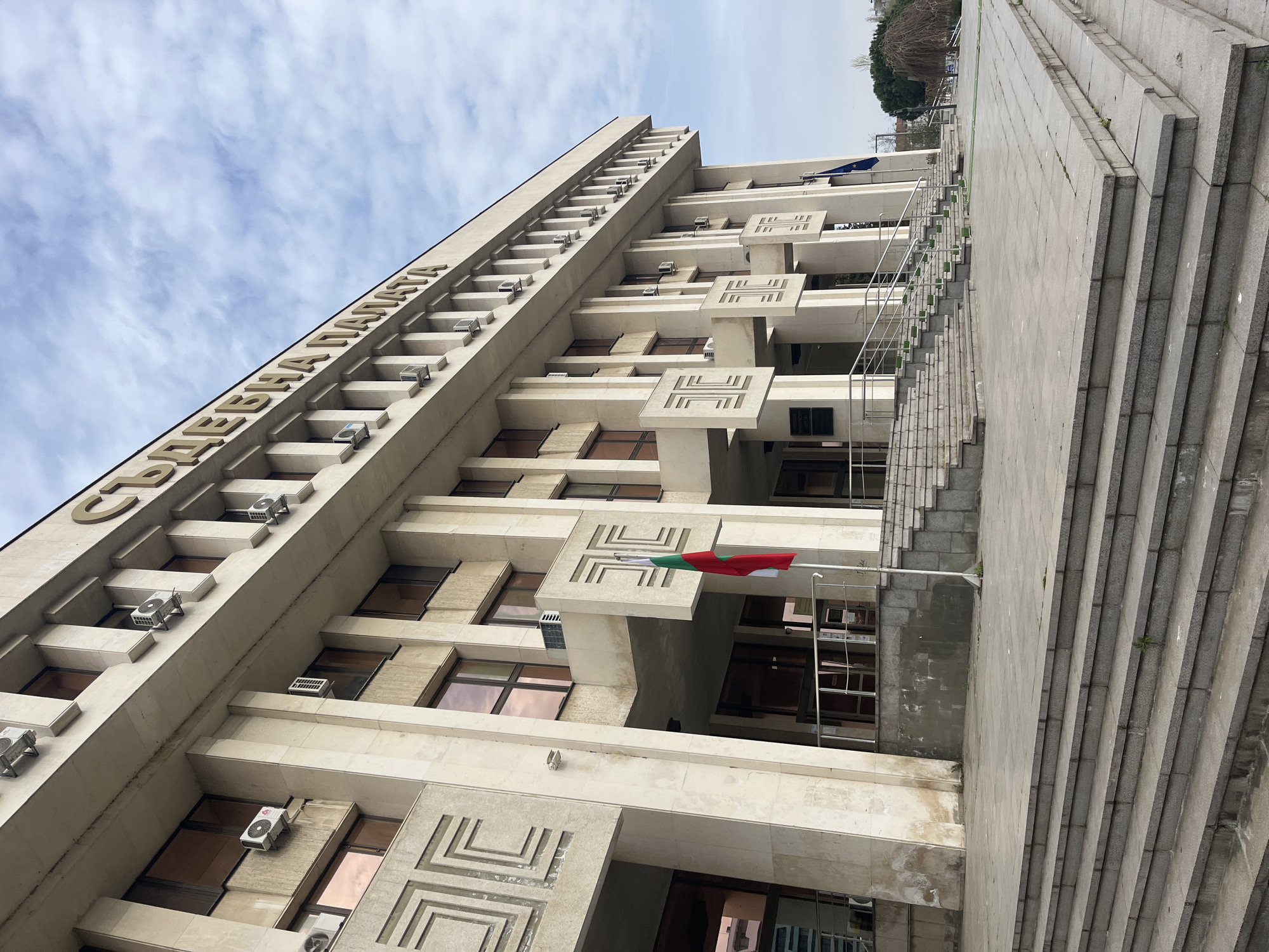 The image depicts the Burgas Court House in Burgas, Bulgaria. The low-angle shot showcases the building's height and brutalist architectural style. The facade is made of light-colored stone or concrete with decorative geometric patterns and rows of windows, each fitted with an air conditioning unit. The name of the court is written in large letters along the top of the building. A Bulgarian flag is displayed on a pole extending from one of the upper floors. To the right, a set of wide concrete steps leads up to the building's entrance. The sky above is partly cloudy.