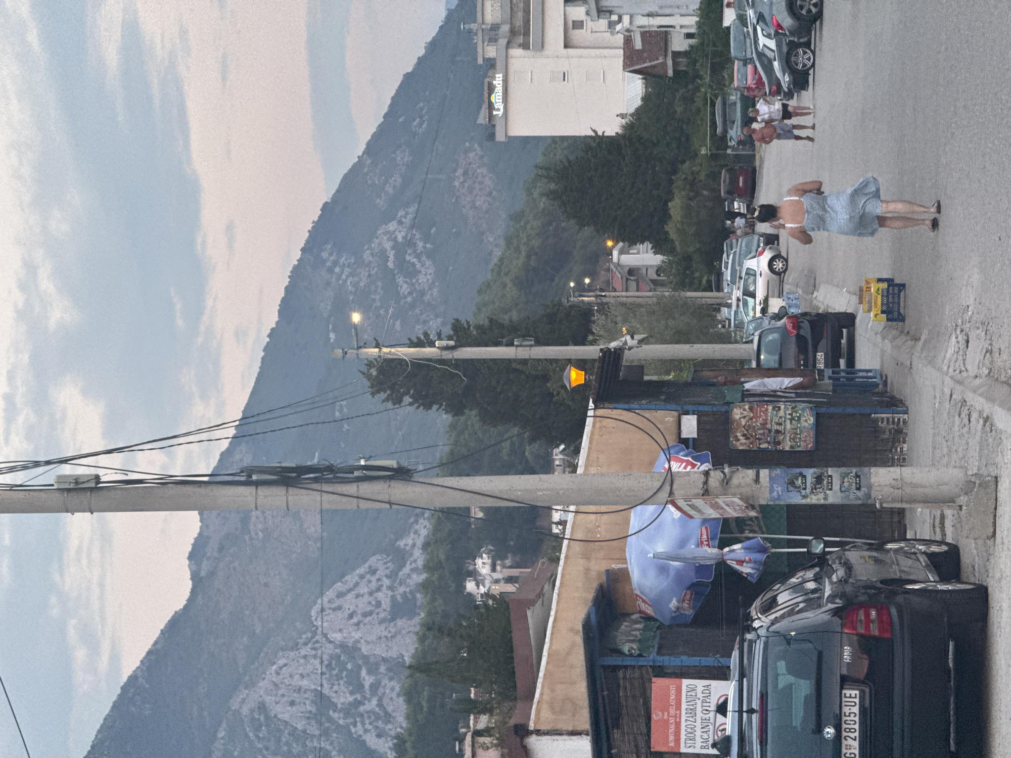 In the city of Sutomore, Montenegro, on an overcast day, a street scene unfolds with a prominent utility pole in the foreground, supporting a network of wires and small electrical boxes. To the left, a black station wagon is parked. Behind the car, a small, open-fronted building with a blue awning is visible, displaying a red sign with white lettering that reads "KOMUNALNE DJELATNOSTI STROGO ZABRANJENO BACANJE OTPADA," indicating a prohibition against littering. A blue umbrella, branded with "Frikon," is propped open near the building.
Further down the street, various cars are parked along the roadside. In the mid-ground, a white building with the "Lamadu" logo illuminated in green and yellow is situated. A woman in a light blue dress walks away from the camera, her dress flowing around her. In the background, a range of tree-covered mountains forms a dramatic backdrop under a pale sky with scattered clouds. The overall atmosphere is that of a moderately populated, developing coastal town.