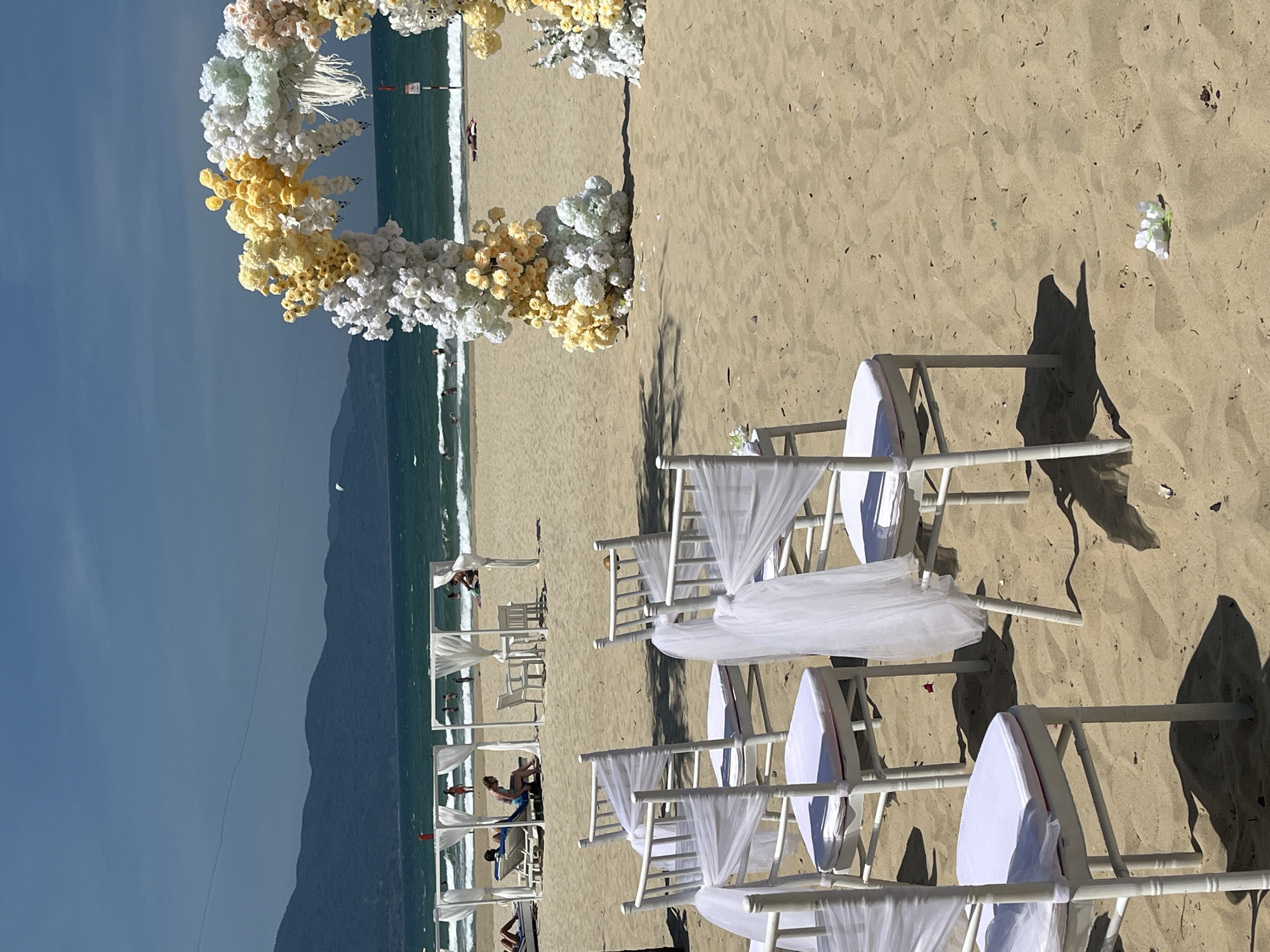 The image depicts a serene beach setting prepared for a special event, likely a wedding, in Đà Nẵng, Vietnam. A prominent semi-circular arch, lavishly adorned with white and yellow artificial flowers, stands on the light brown sand, facing the ocean.
In front of the floral arch, several rows of white chairs with white sheer fabric drapes and white seat cushions are meticulously arranged on the sand, forming an aisle. Long shadows cast by these chairs and other elements suggest either early morning or late afternoon sun under a clear blue sky. Small white flowers are scattered on the sand around the event area.
Beyond the immediate setup, the blue-green waves of the ocean gently meet the shore. Along the beachfront, several white, open-sided beach cabanas or tents are visible, with a few individuals relaxing or engaged in activities near the water. In the far background, a range of dark mountains stretches across the horizon. A small red sign or buoy is faintly visible in the distant ocean, but any text on it is unreadable. The overall scene conveys a sense of romantic tranquility and natural beauty, highlighting the blend of celebration with the coastal environment.