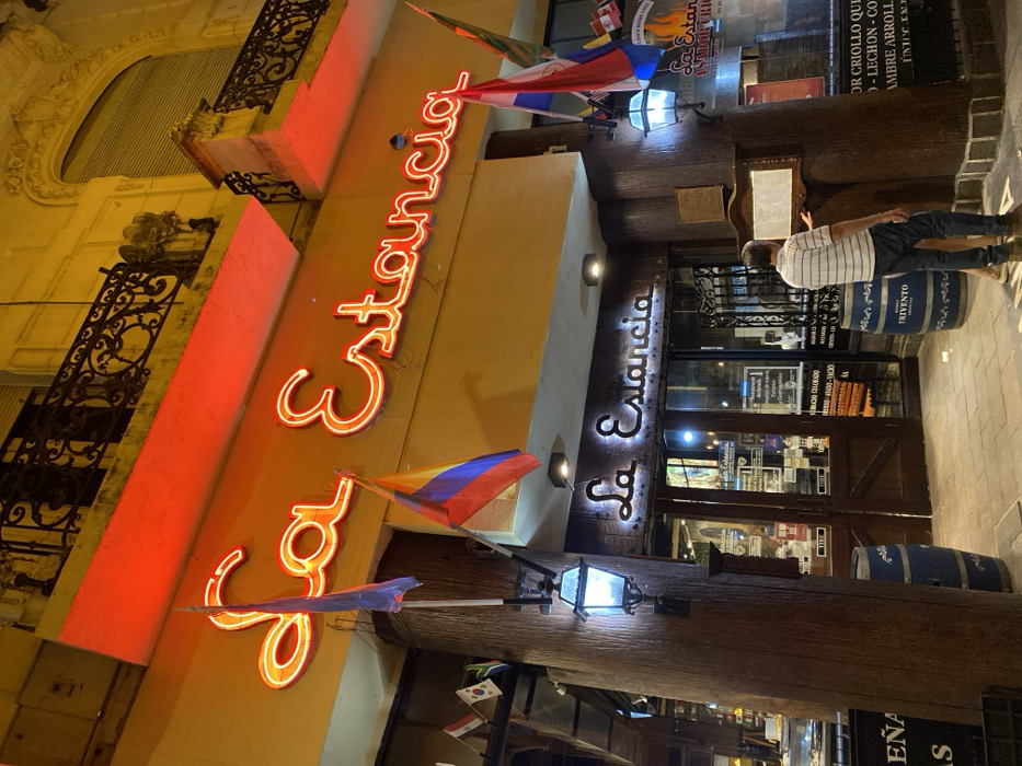 This outdoor nighttime image depicts a restaurant named "La Estancia." The bright neon signage in a flowing script is visible on the upper part of the building façade. Below, a darker, illuminated sign marks the entrance. Country flags adorn the space. A man stands near the entrance, seemingly reviewing a menu or sign. Decorative barrels are placed near the entrance. Architectural details, including ornate ironwork on balconies, add to the building's character.