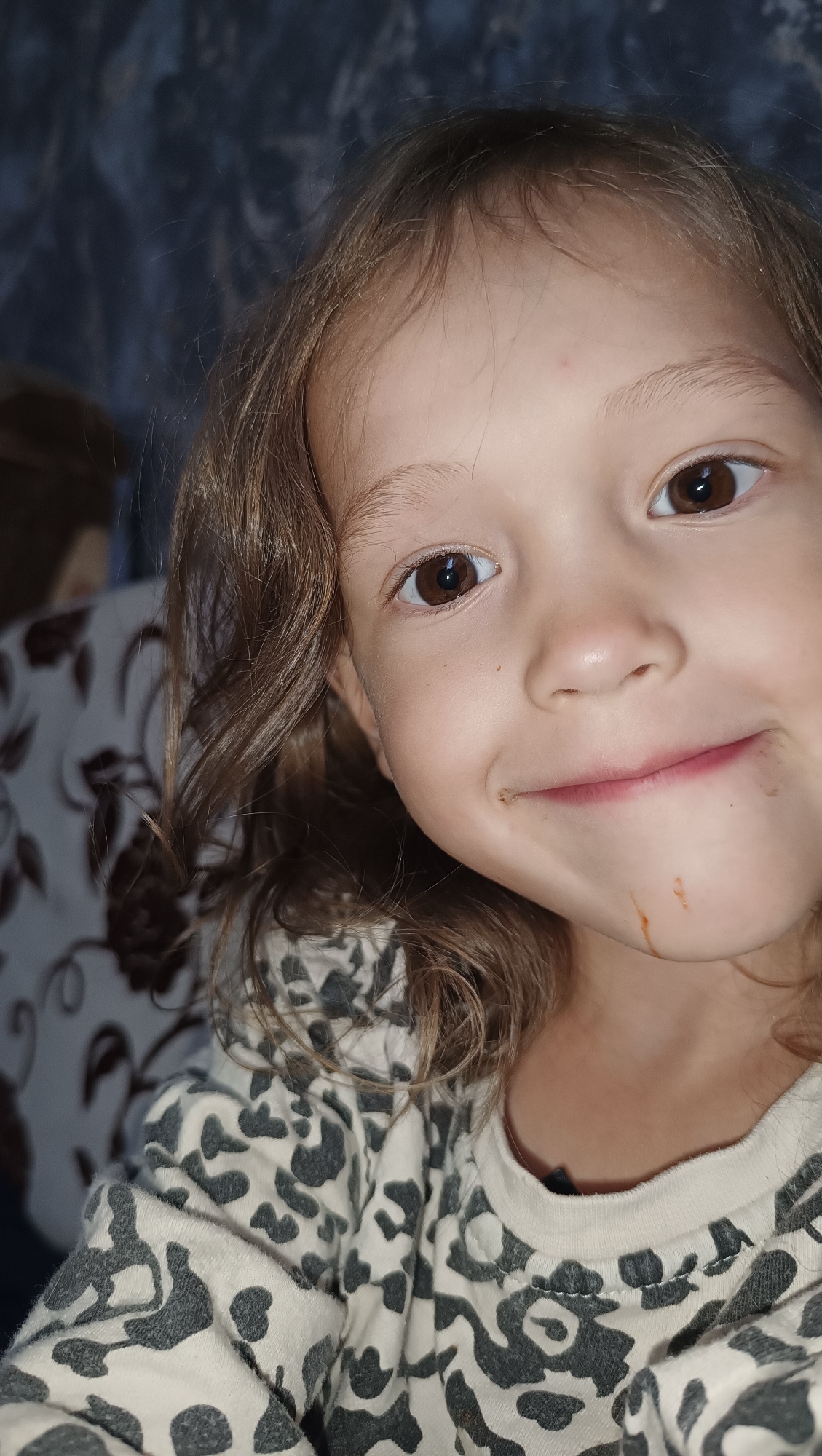A close-up, eye-level, indoor portrait shows a young child with light brown, wavy hair and brown eyes. The child's head is turned slightly to the right, with their gaze directed towards the camera. They have a slight smile, and there appears to be some brown staining on their chin and around their mouth, suggesting they may have recently eaten something messy, possibly chocolate. The child is wearing a white and gray patterned long-sleeved shirt. The background is out of focus, showing a dark, textured surface on the left and a floral-patterned fabric on the right. The lighting is dim, creating a slightly shadowy atmosphere. This image was taken in the city of Karsha, Russia.