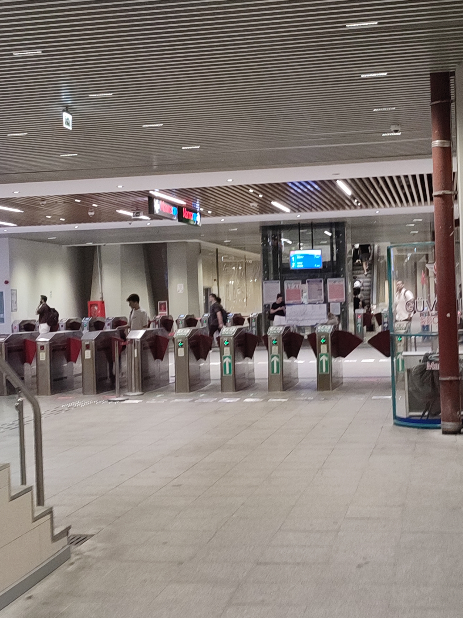 The image depicts the interior of a modern transportation hub, likely a metro station, in Kadıköy, Turkey. The environment features a sleek, contemporary design with a metal slatted ceiling and tiled flooring. In the foreground, a series of automated turnstiles are arranged in a row, with some people in the process of passing through them. The turnstiles are metallic with red and green indicator lights, and some have clear plastic barriers.
Several individuals are visible. To the left, a person wearing a dark jacket and a backpack is seen interacting with a turnstile. Further back, another person in a light-colored shirt and cap is also at a turnstile. In the mid-ground, a group of people are heading towards an escalator, suggesting an upward movement within the station. On the right side of the frame, a glass-enclosed structure is partially visible, containing a trash receptacle.
Overhead, directional signs with red and white lettering are mounted, indicating destinations. A digital display screen showing a blue interface is visible in the background. The lighting in the station is a mix of recessed fixtures in the ceiling and illuminated signs. The overall atmosphere suggests a busy, functional public space.